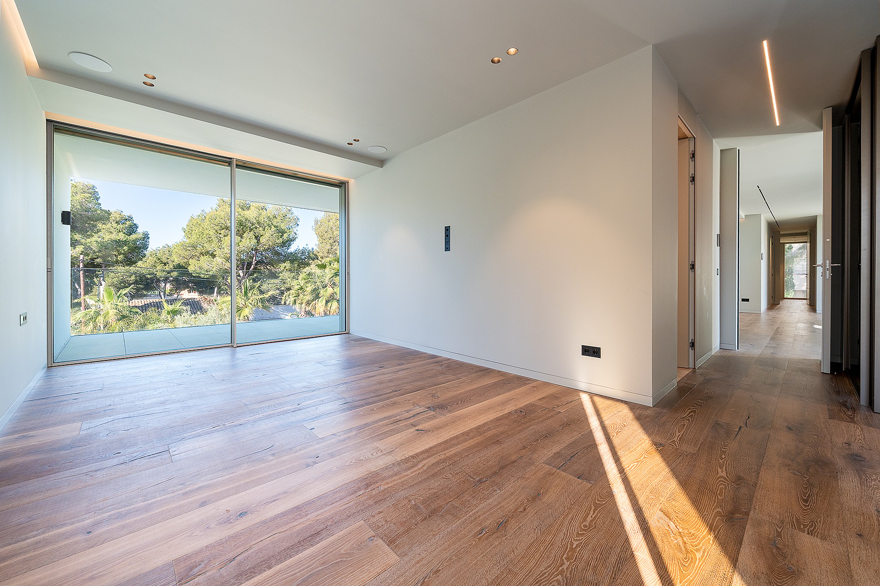 Empty modern bedroom with wide oak floorboards, white walls, and a large sliding glass door looking out toward green trees.
