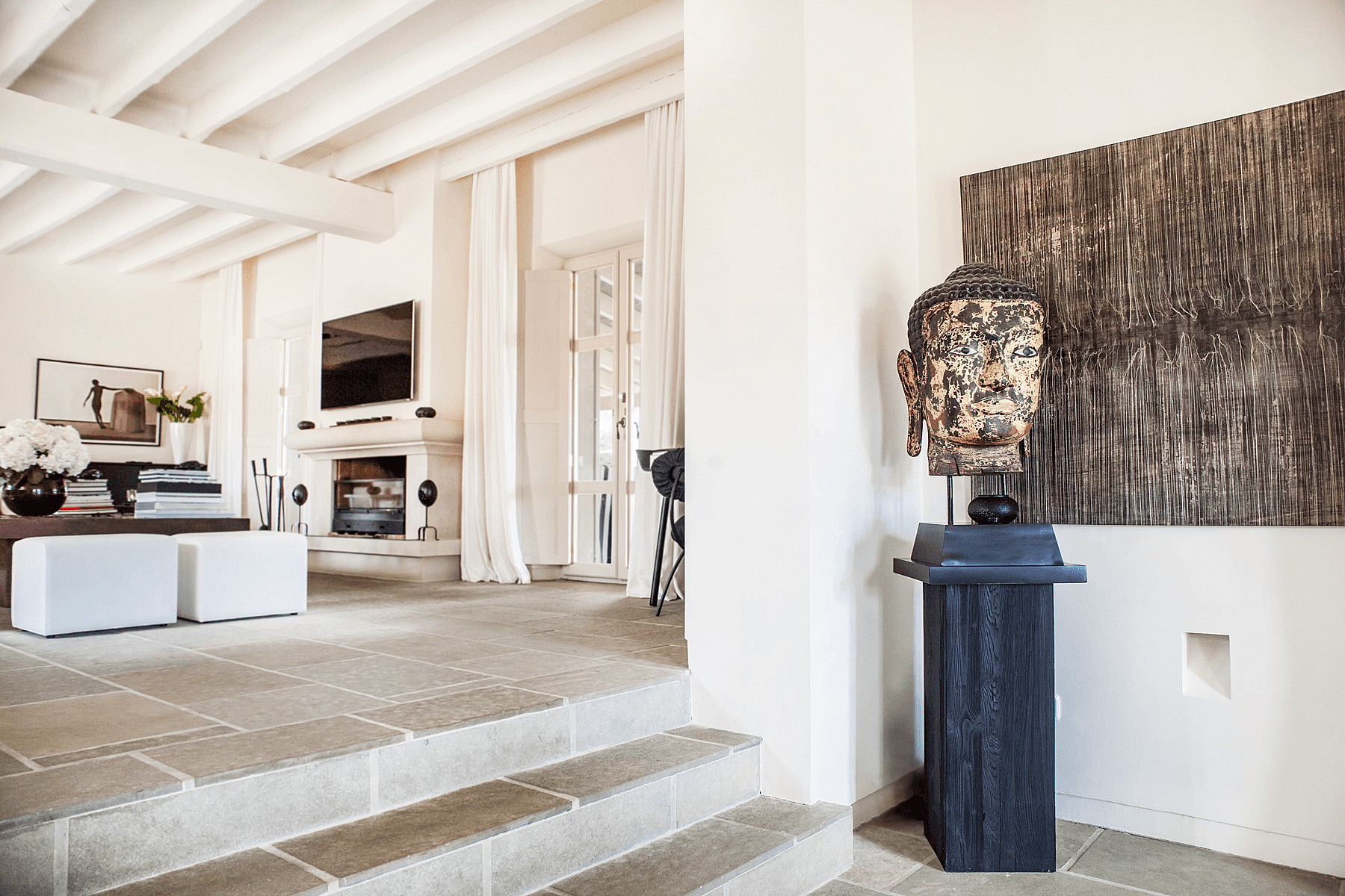 A living area with a stone-paved sunken floor, exposed white ceiling beams, and a fireplace, featuring a sculpture on a pedestal.
