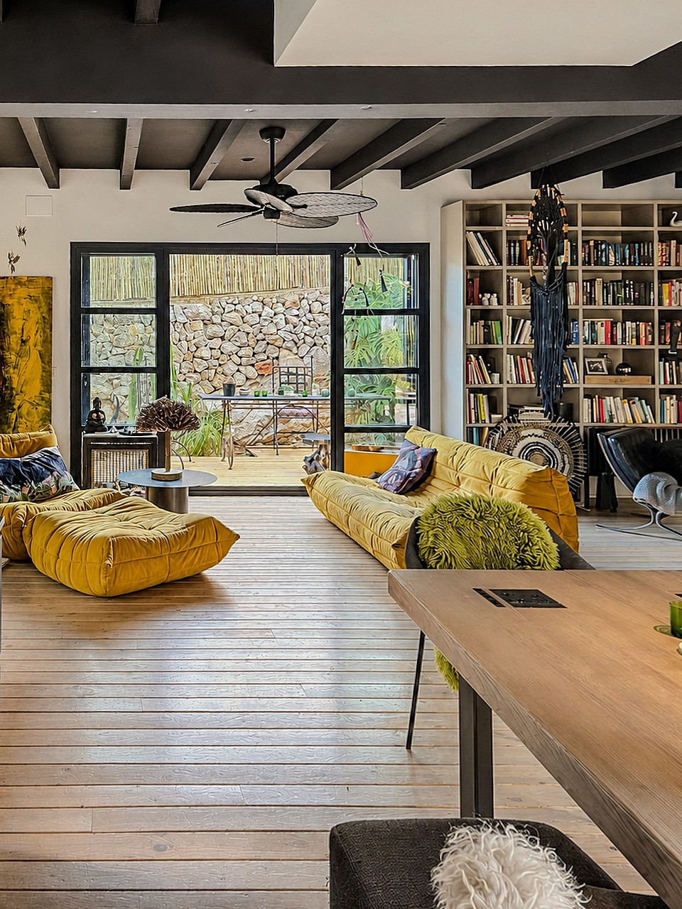 A living area with light wood floors, a large yellow sofa, and a wall-to-wall integrated bookshelf opening onto a stone terrace.