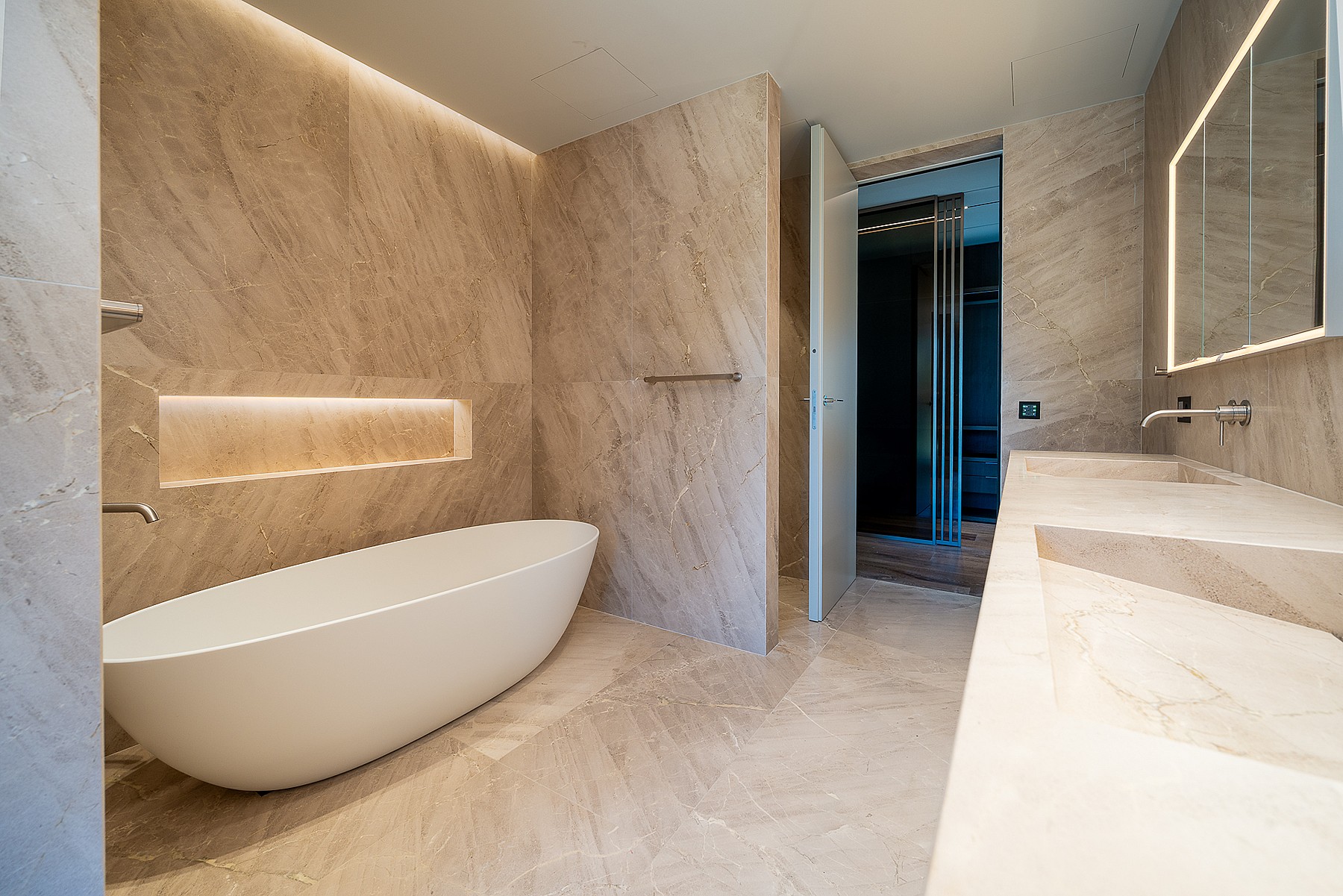 Modern master bathroom featuring a white oval freestanding bathtub against beige stone walls, a double vanity, and recessed LED lighting.