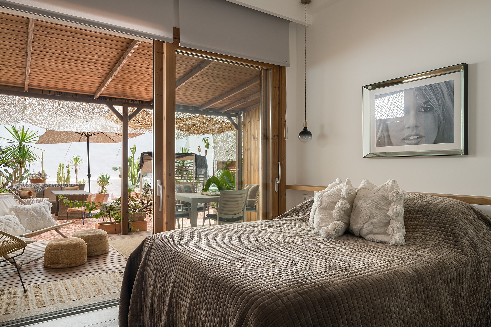 A bedroom with a textured brown bedspread, opening through large sliding glass doors to a furnished plant-filled terrace.
