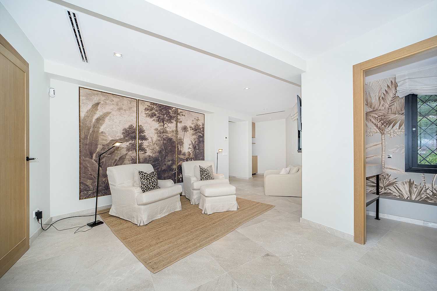 Interior room featuring beige armchairs on a jute rug, a large forest-motif wall mural, and light stone flooring.