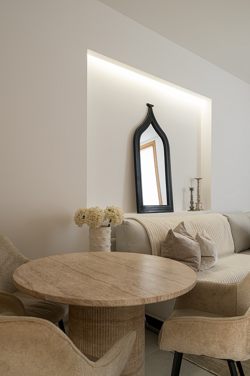 A round travertine stone dining table set in a living room with a beige textured sofa and a black arched mirror on the wall.