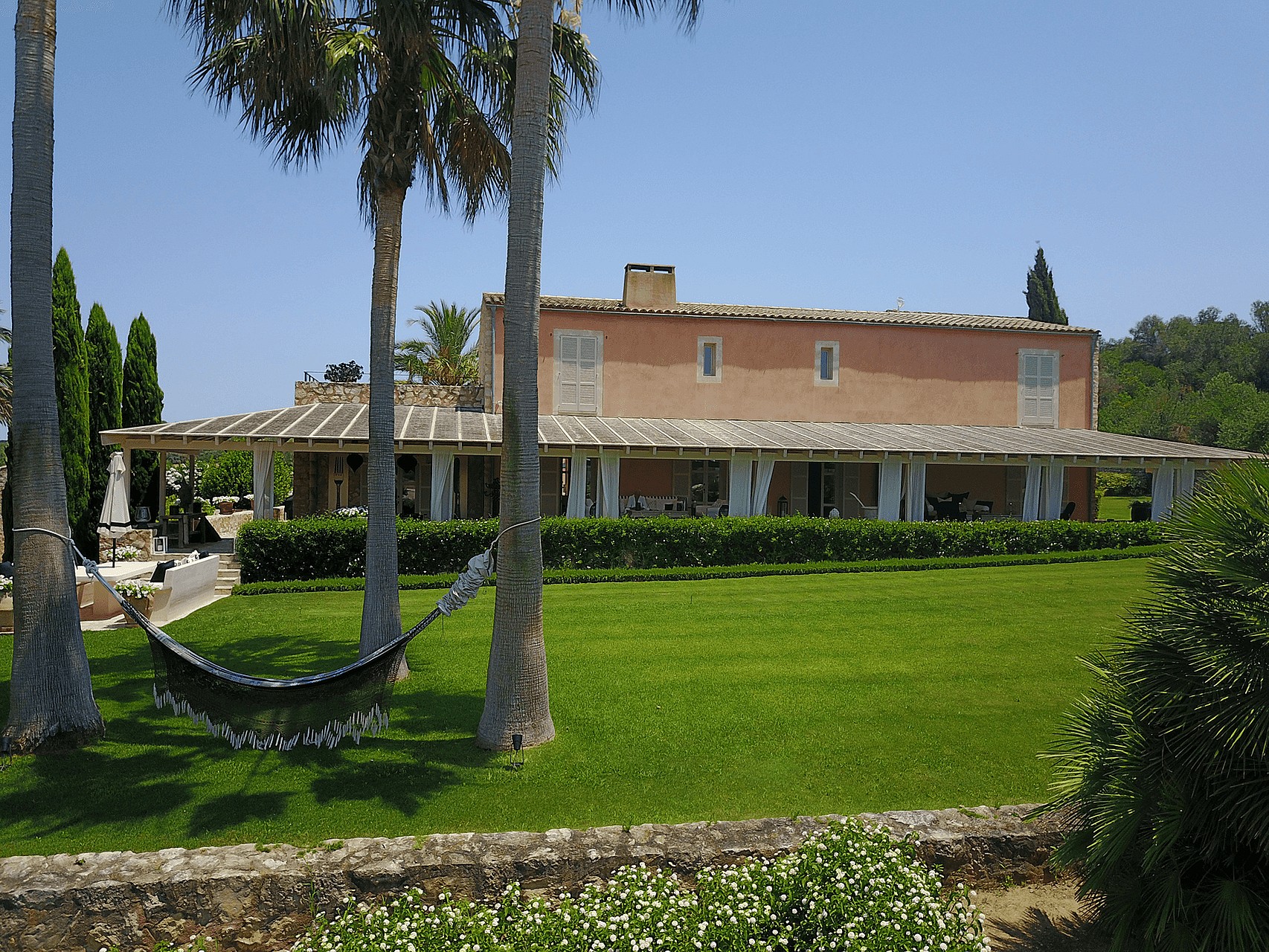 Exterior view of a traditional terracotta-toned finca with a long covered porch, palm trees, and a hammock on a green lawn.