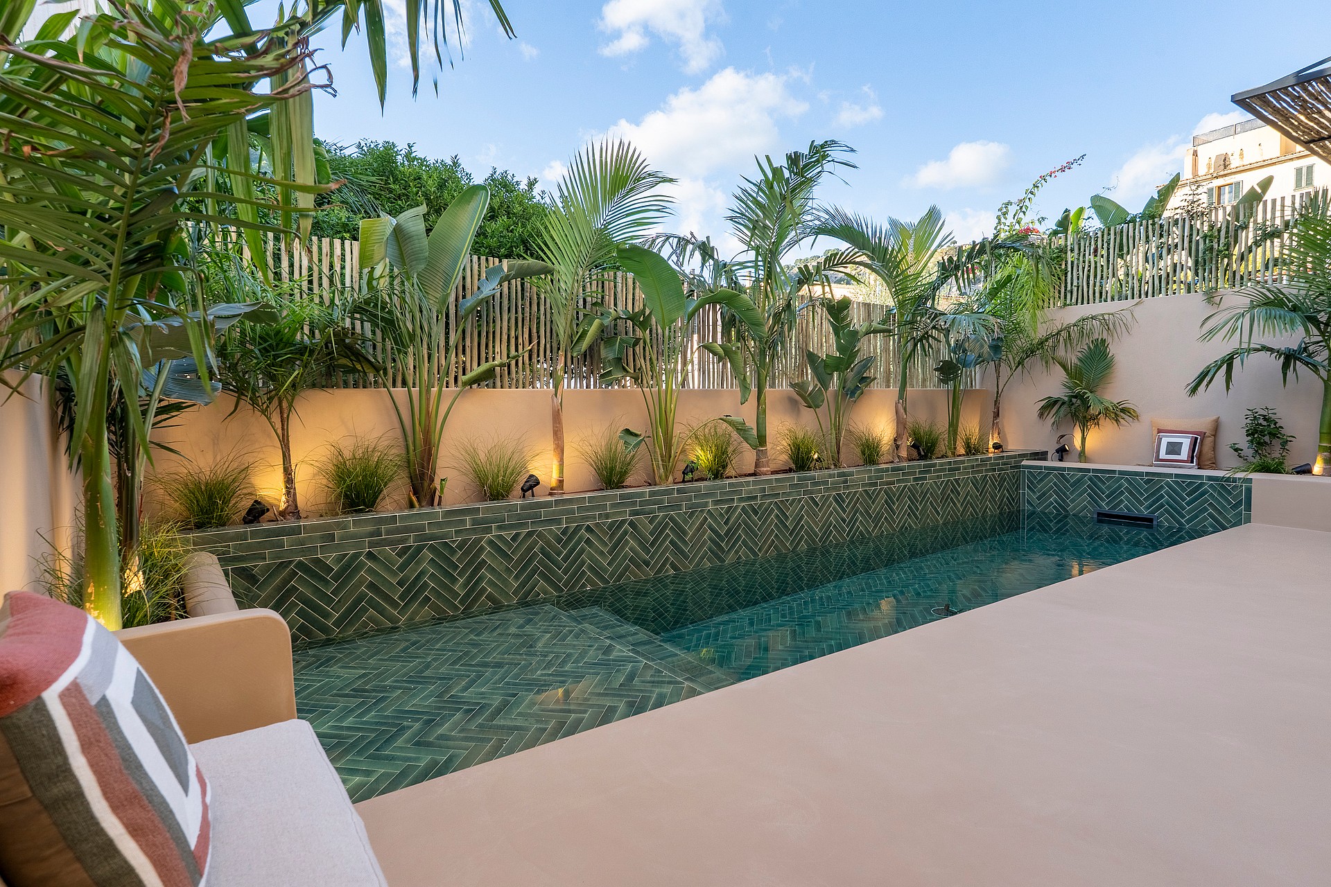 A narrow swimming pool with green patterned tiles is surrounded by a beige stone terrace and lush palm trees.