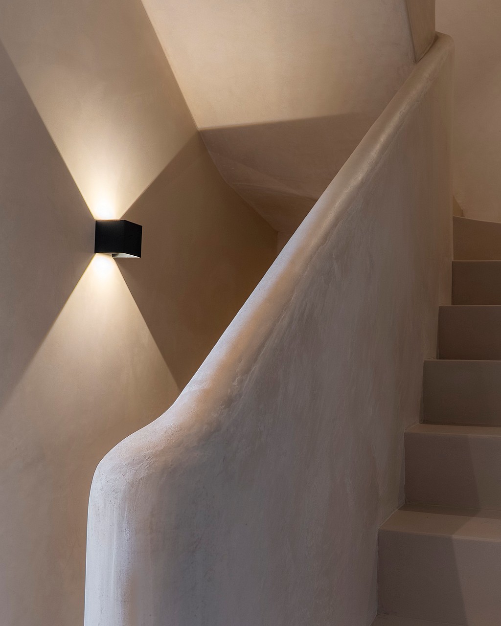 A staircase with off-white plaster walls and a textured balustrade, illuminated by a sharp geometric wall light.