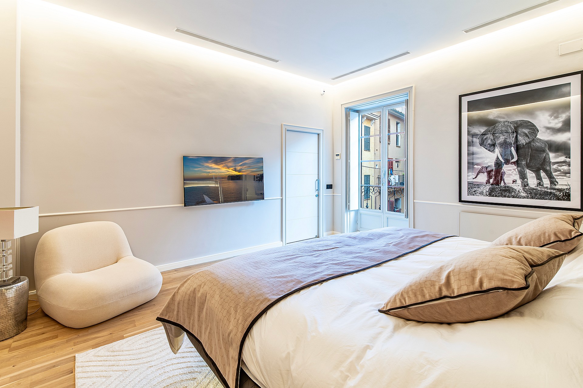 A modern bedroom featuring a king-sized bed, cream-colored armchair, recessed cove lighting, and a view of an Old Town street.