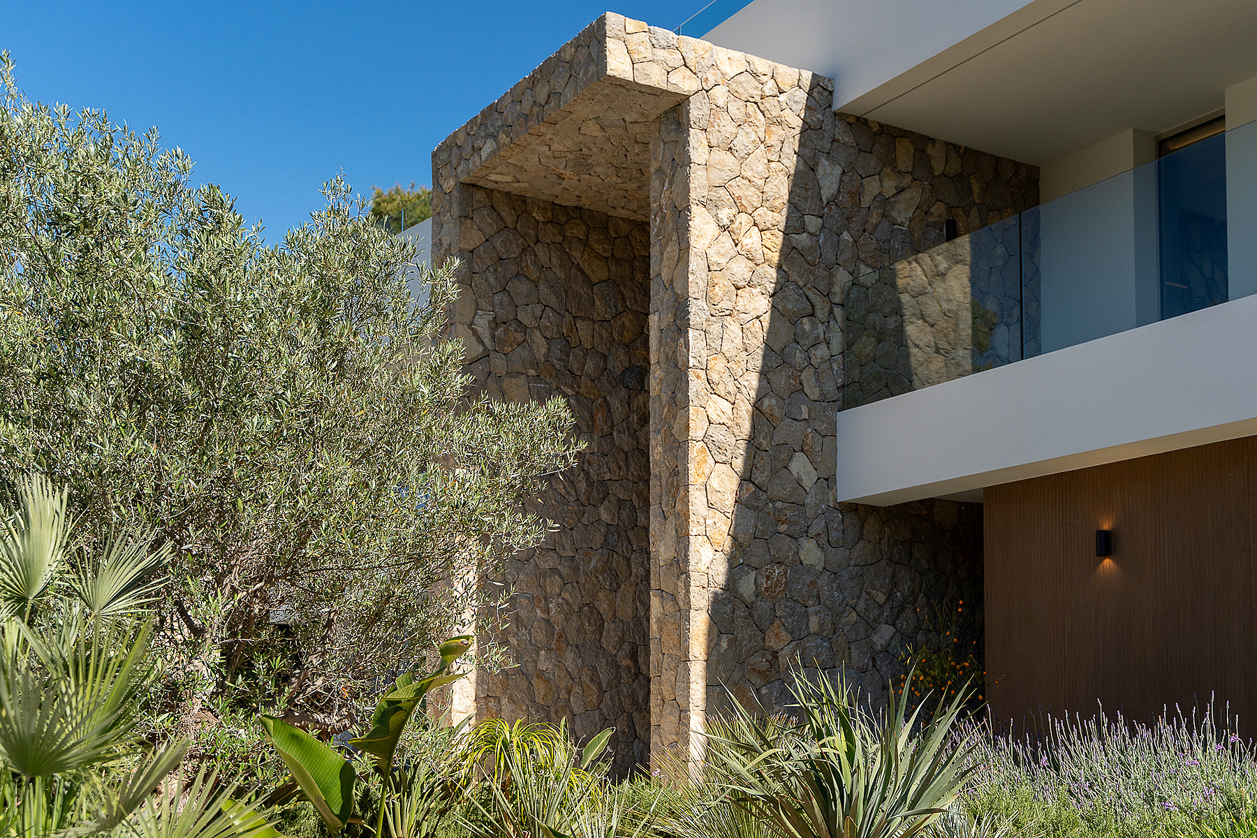 Close up of a modern villa exterior showing dry stone wall architecture, an olive tree, and a glass balcony railing.