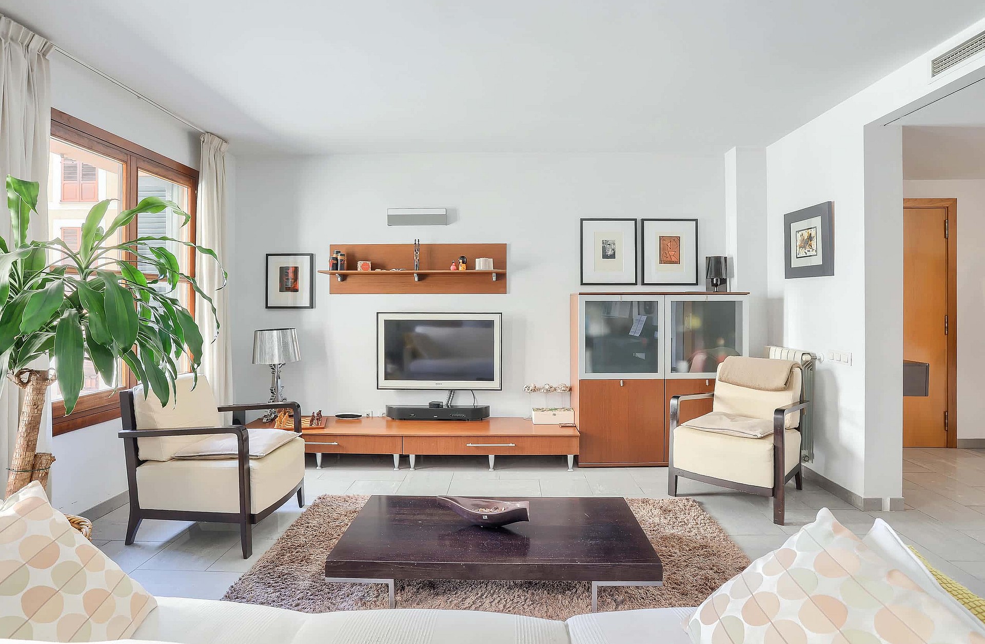 Living room featuring cream armchairs, a low dark wood coffee table, and a wall-mounted television unit on white walls.