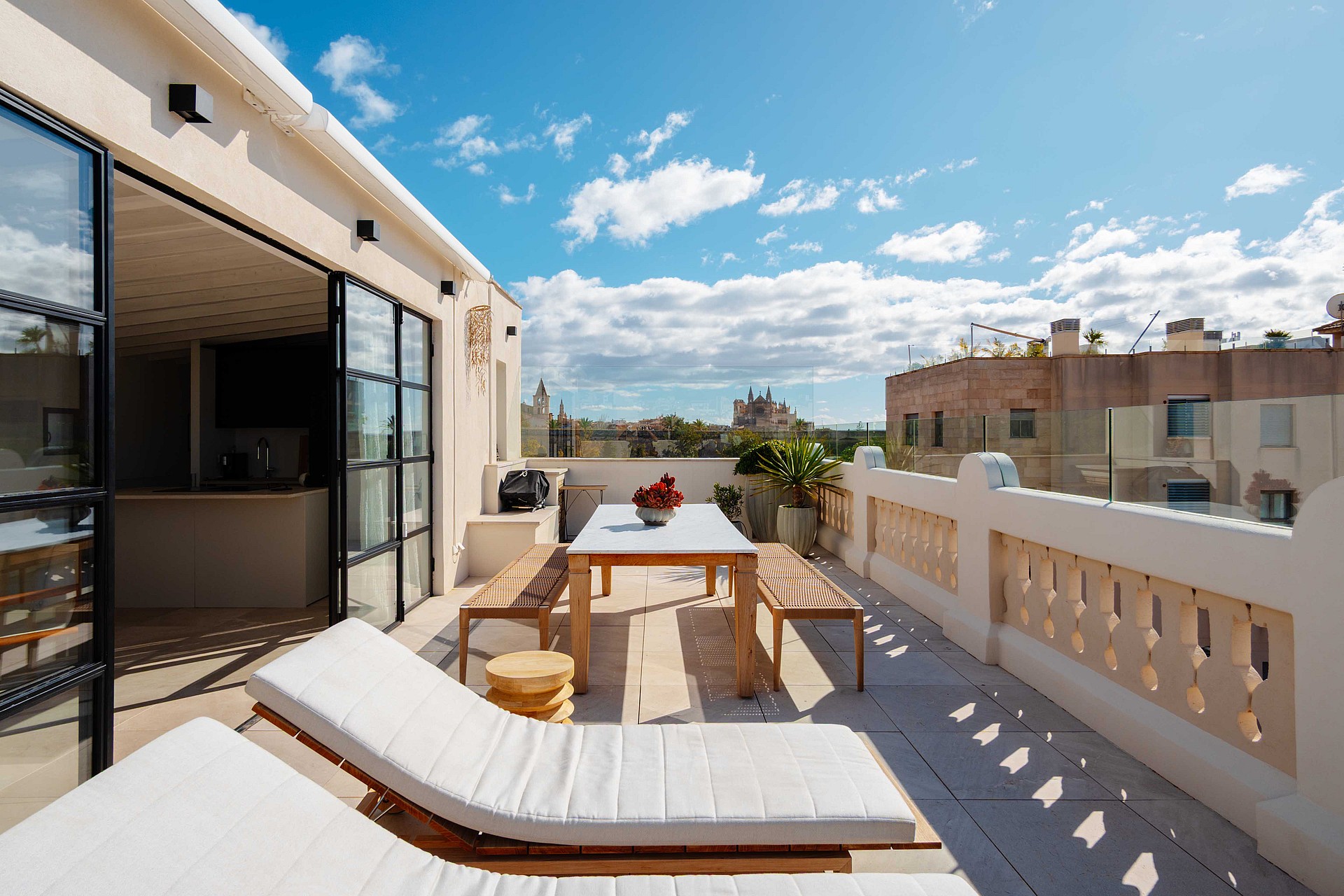 A sunny rooftop terrace in Santa Catalina with white sun loungers, a wooden dining table and benches, and iron-framed glass doors.