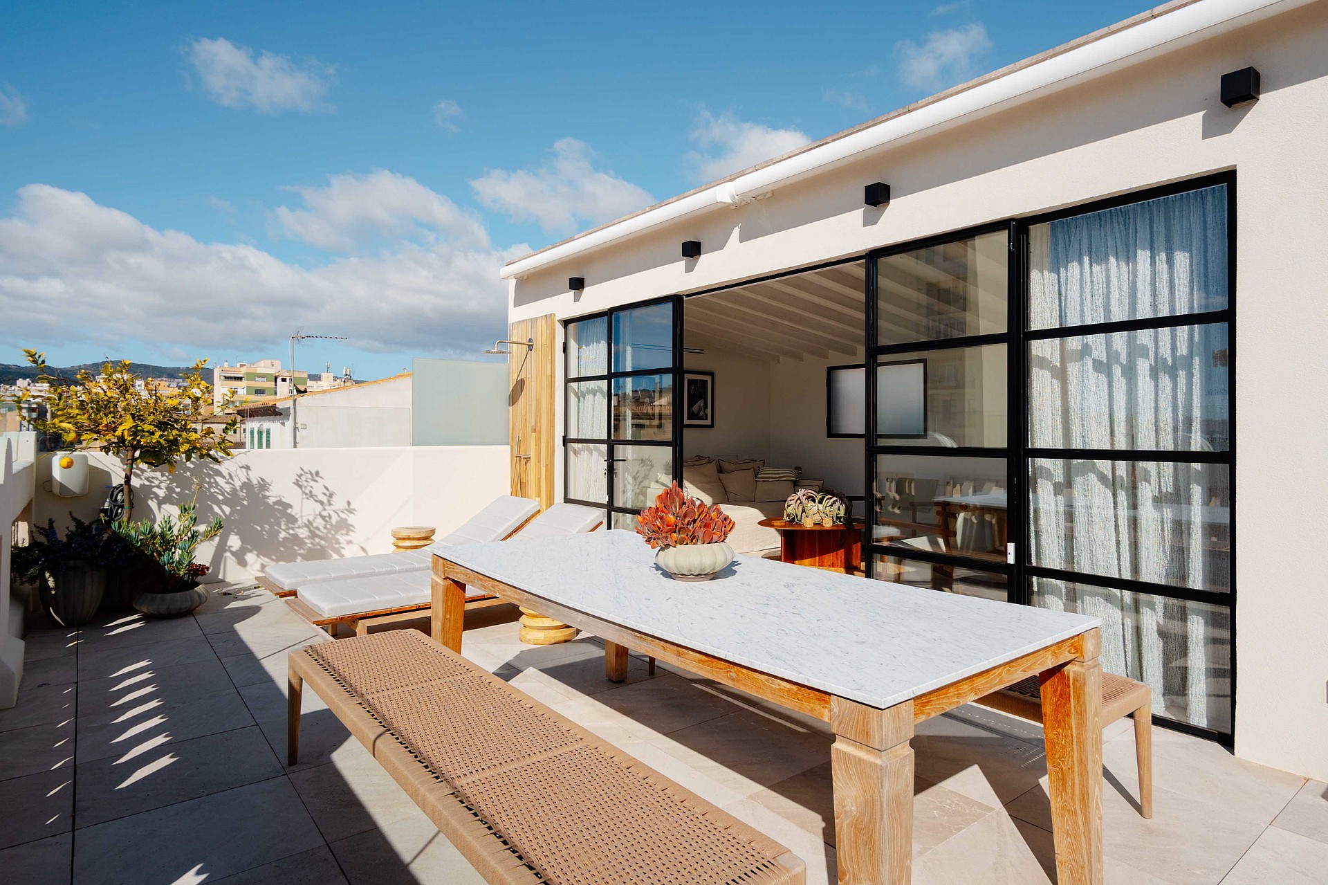 A rooftop terrace featuring a wooden dining table and benches, connected to an indoor living area by black-framed glass doors.
