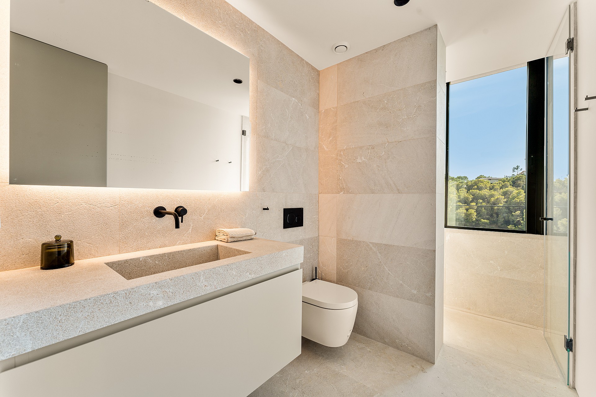 A minimalist bathroom featuring large-format beige tiles, a wall-hung toilet, a floating vanity with an integrated mirror, and a glass-enclosed shower area with views of greenery.