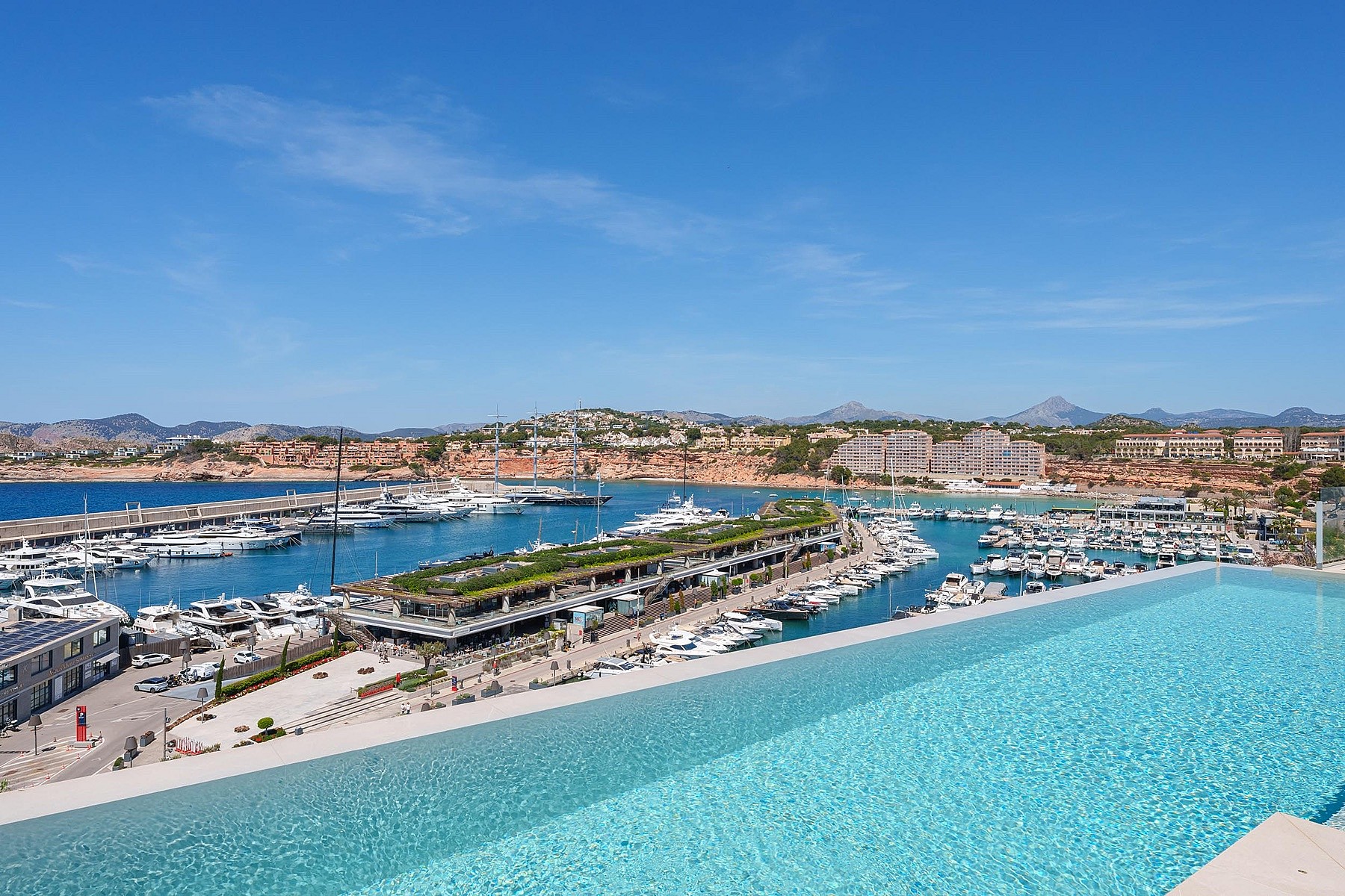 An elevated infinity pool looking down over a marina filled with yachts, with a coastal landscape in the background.