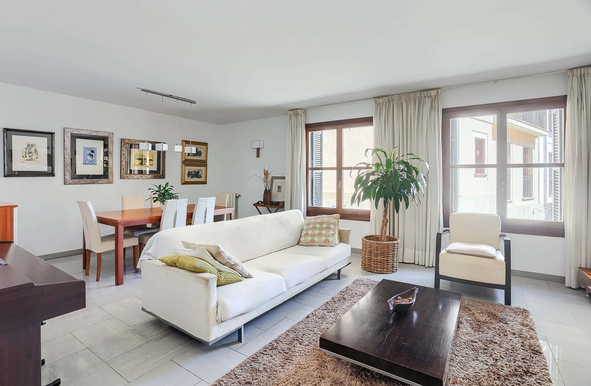 A bright living room with a white sofa, dark wooden coffee table, and a dining table for six, featuring neutral decor.