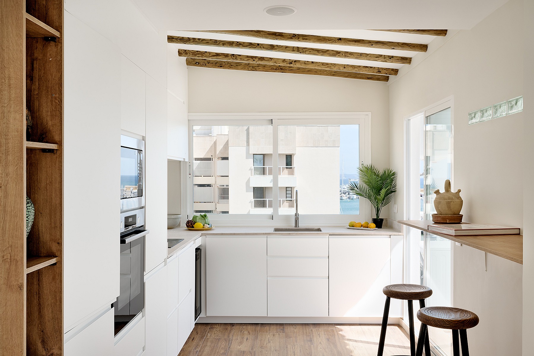 Modern kitchen with white cabinets, wood ceiling beams, and a window overlooking an urban exterior.