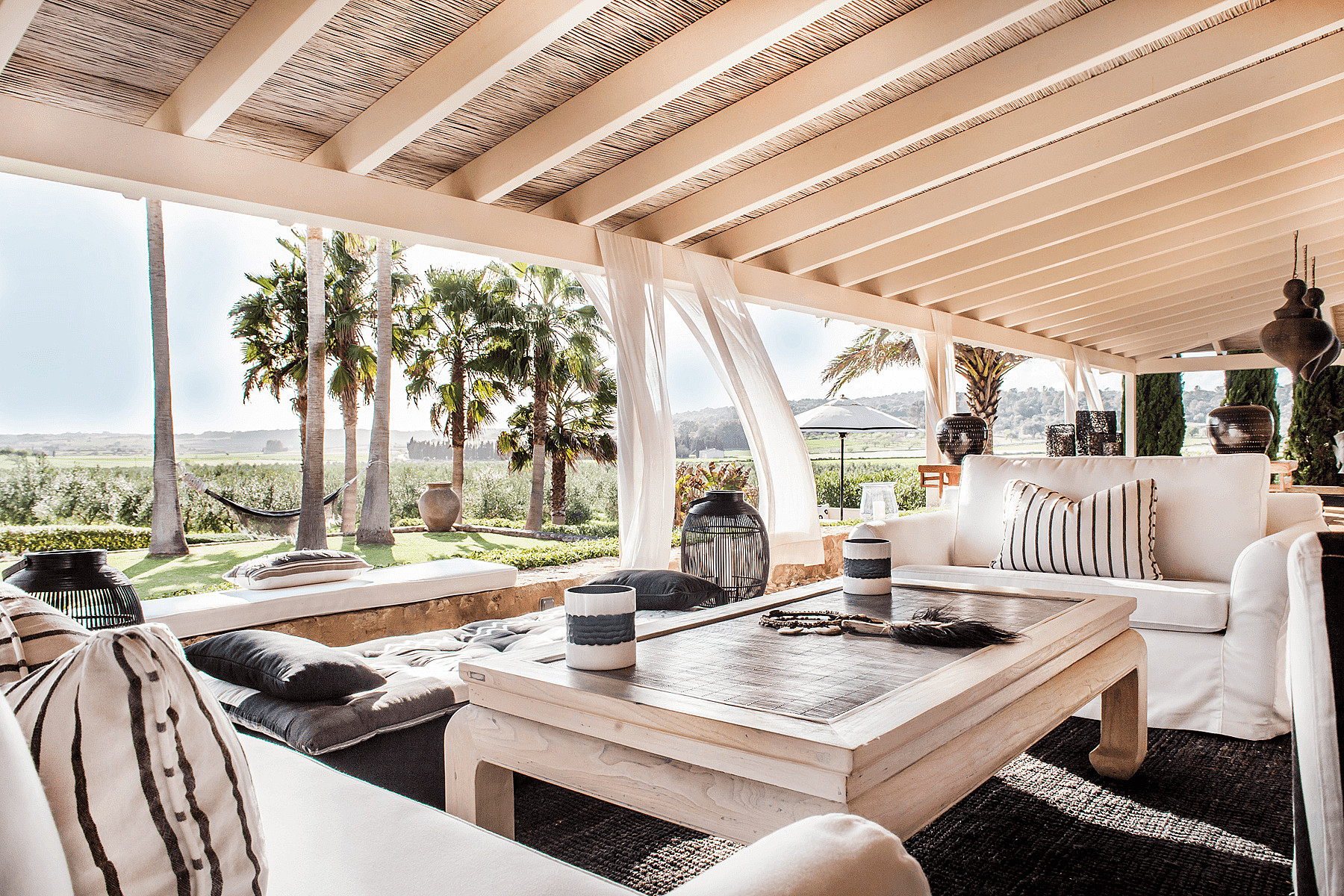 A wide, covered outdoor seating area with white fabric sofas and a wooden coffee table, overlooking a green landscape with palms.
