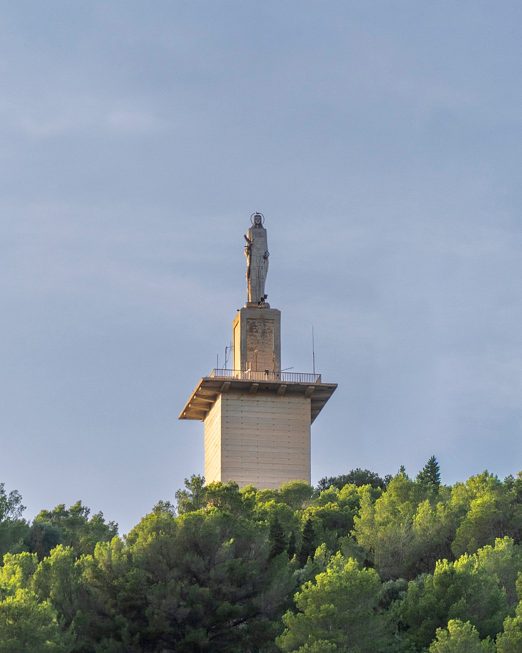 A stone statue on a pedestal positioned atop a lush green hill against a clear sky in Genova.