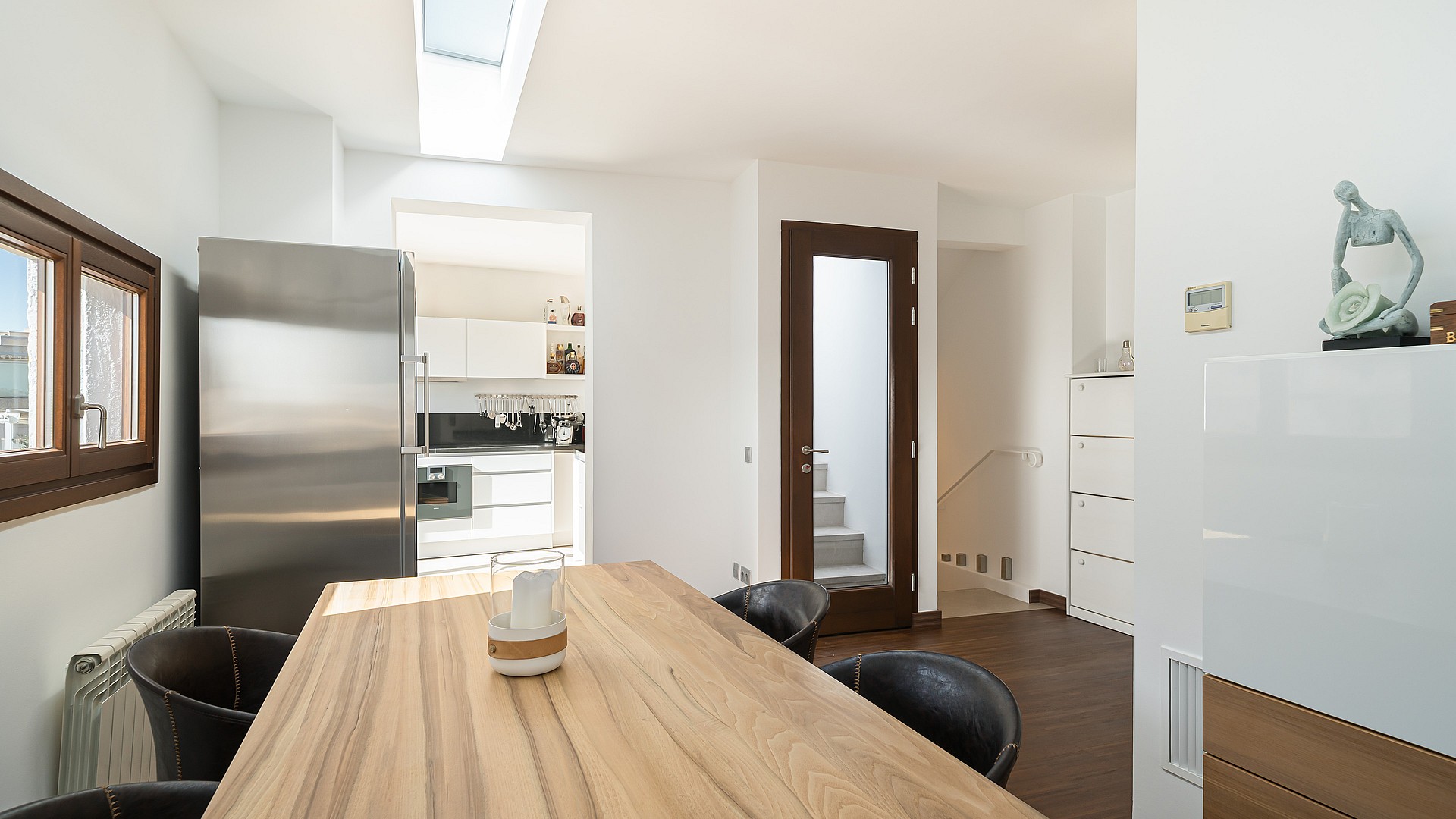 Bright dining space with large oak table, stainless steel fridge, and a skylight in a modern white interior.
