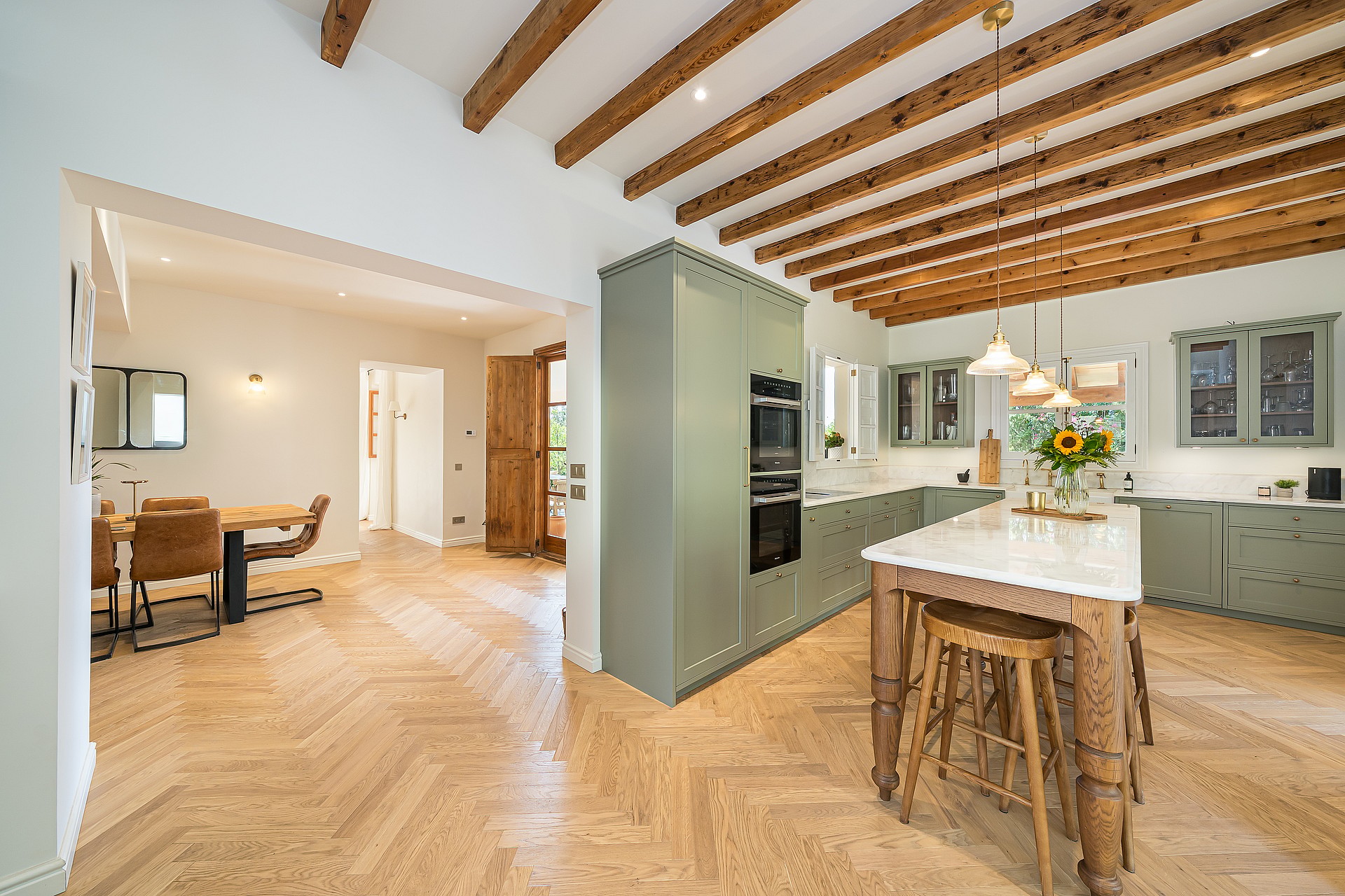 Modern kitchen featuring sage green cabinets, herringbone wood floors, a marble-top island, and exposed timber ceiling beams.