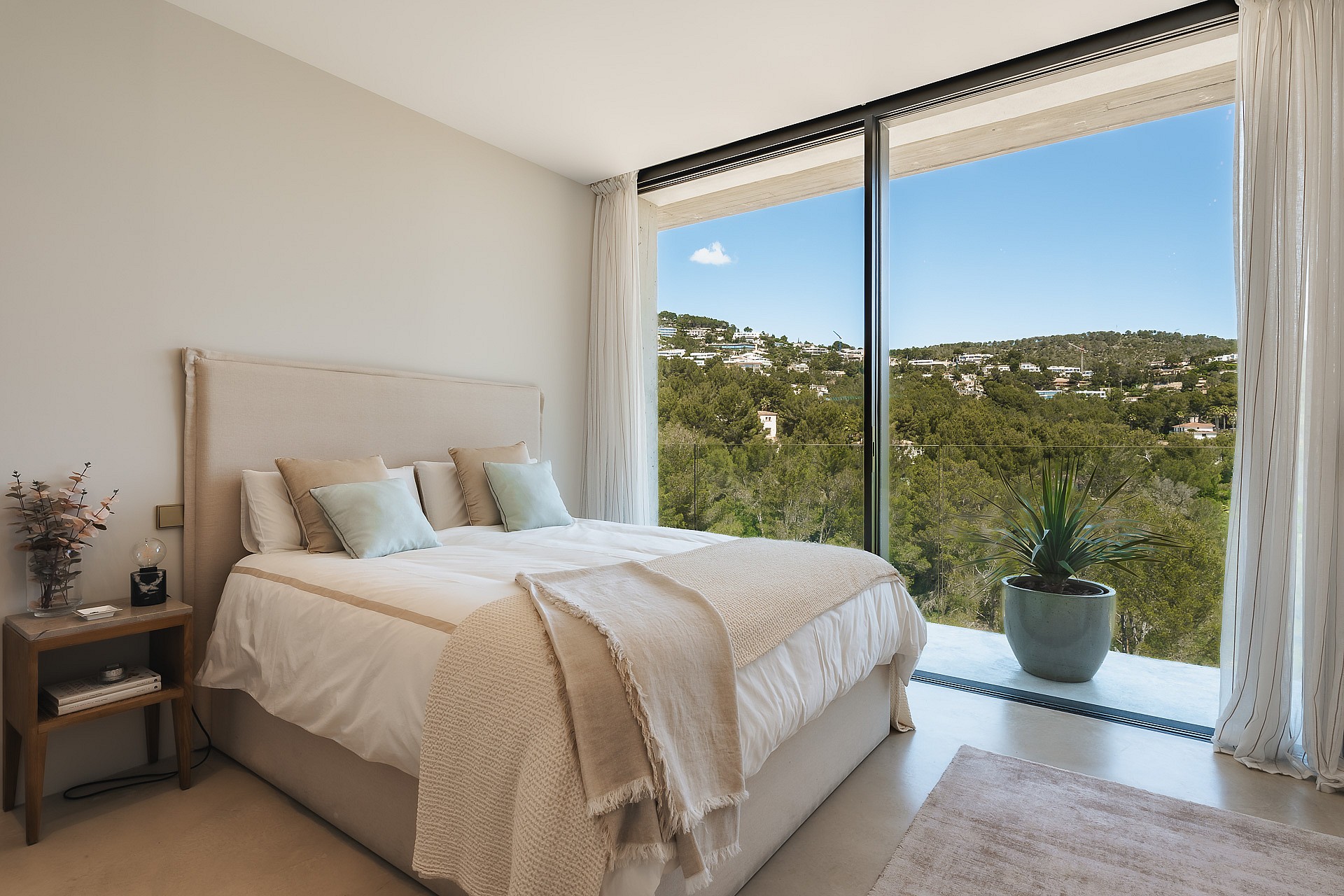 Modern bedroom with floor-to-ceiling windows overlooking green hills, featuring neutral textiles and a wooden side table.