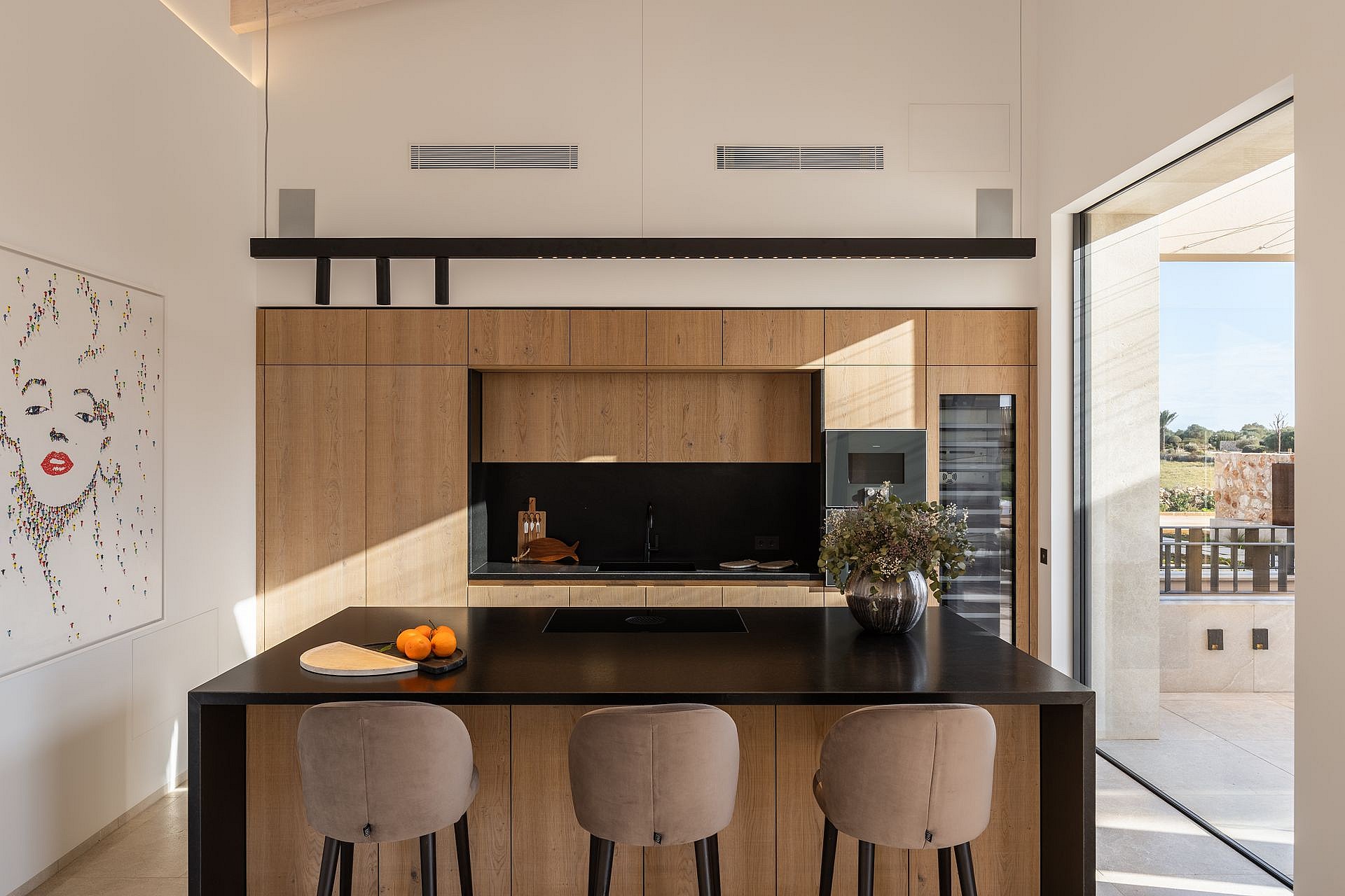 A modern kitchen with wood cabinetry, a black stone island, and three upholstered stools, opening to a sunlit terrace.