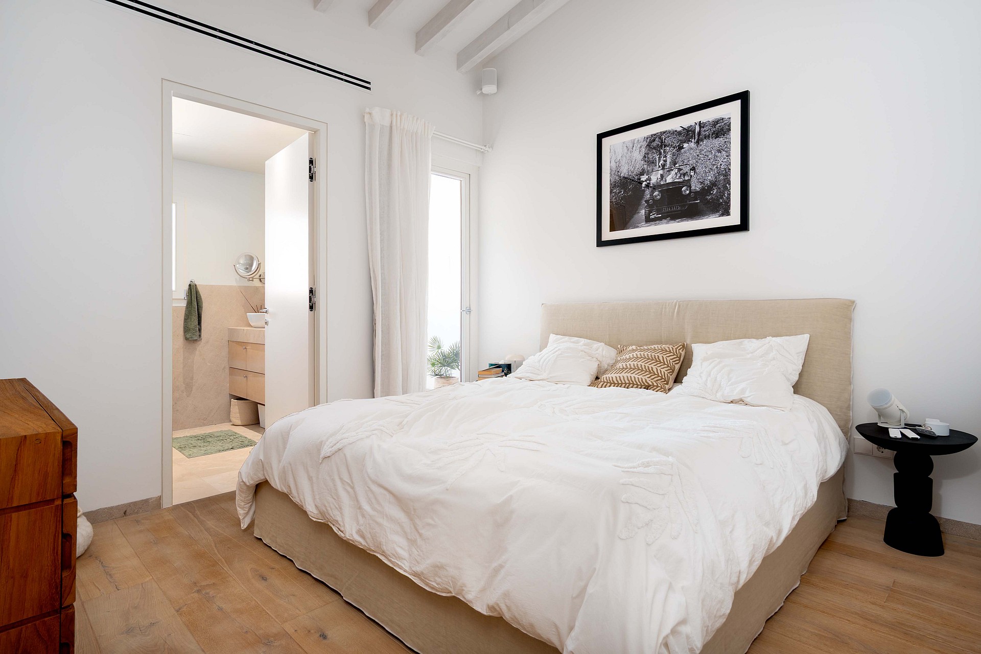 Modern bedroom with a neutral upholstered bed, light oak wood flooring, white walls, and a door leading to a bathroom.