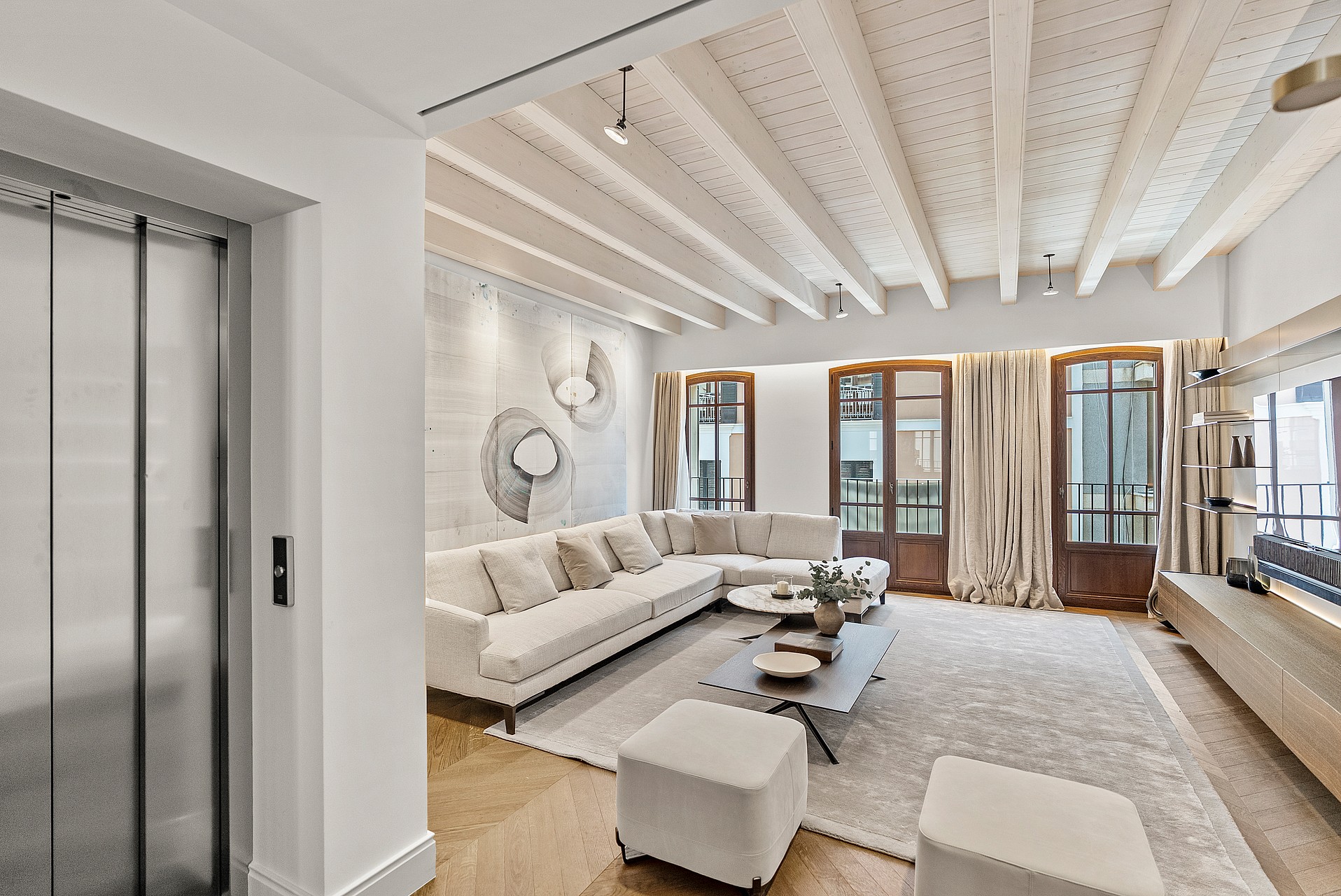 Bright living room with herringbone wood flooring, white sectional sofa, and tall wooden French doors under a beamed ceiling.