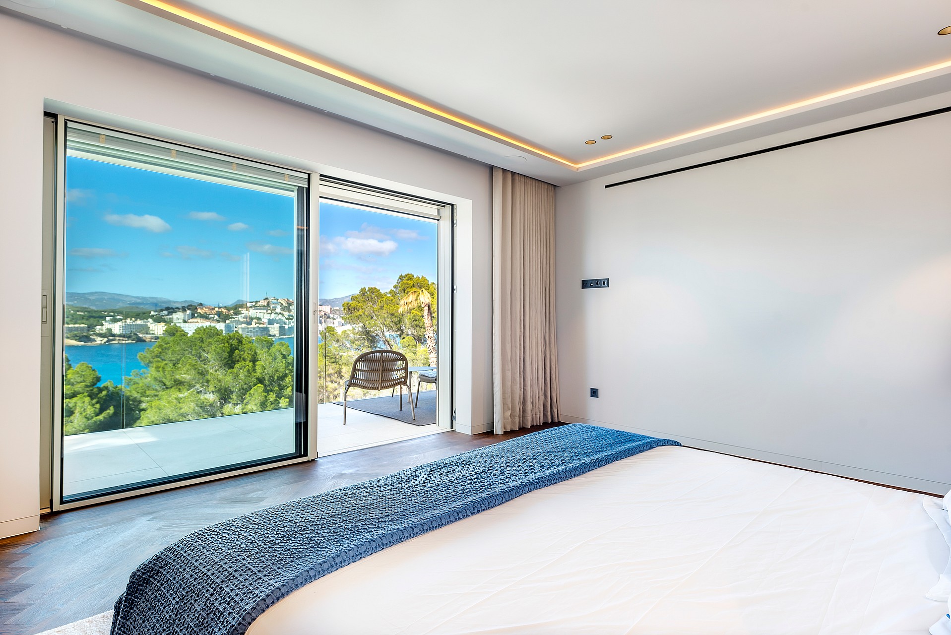 A modern bedroom with floor-to-ceiling sliding glass doors opening onto a terrace overlooking a turquoise coastal bay.