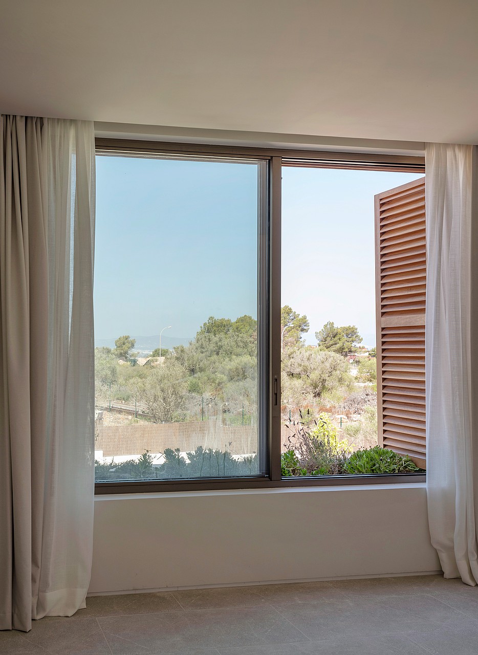 Interior view of a window with brown wooden shutters and white curtains looking out to a sun-drenched green landscape.