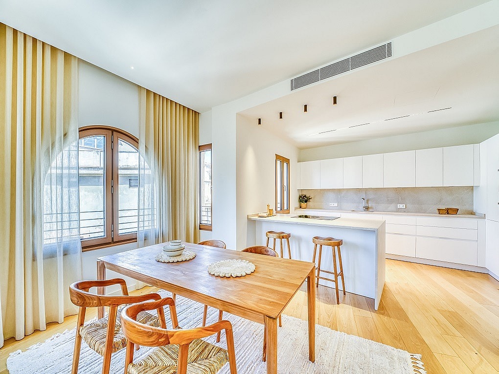 A bright open-plan kitchen and dining room with wooden furniture, white cabinetry, and large arched windows draped in curtains.