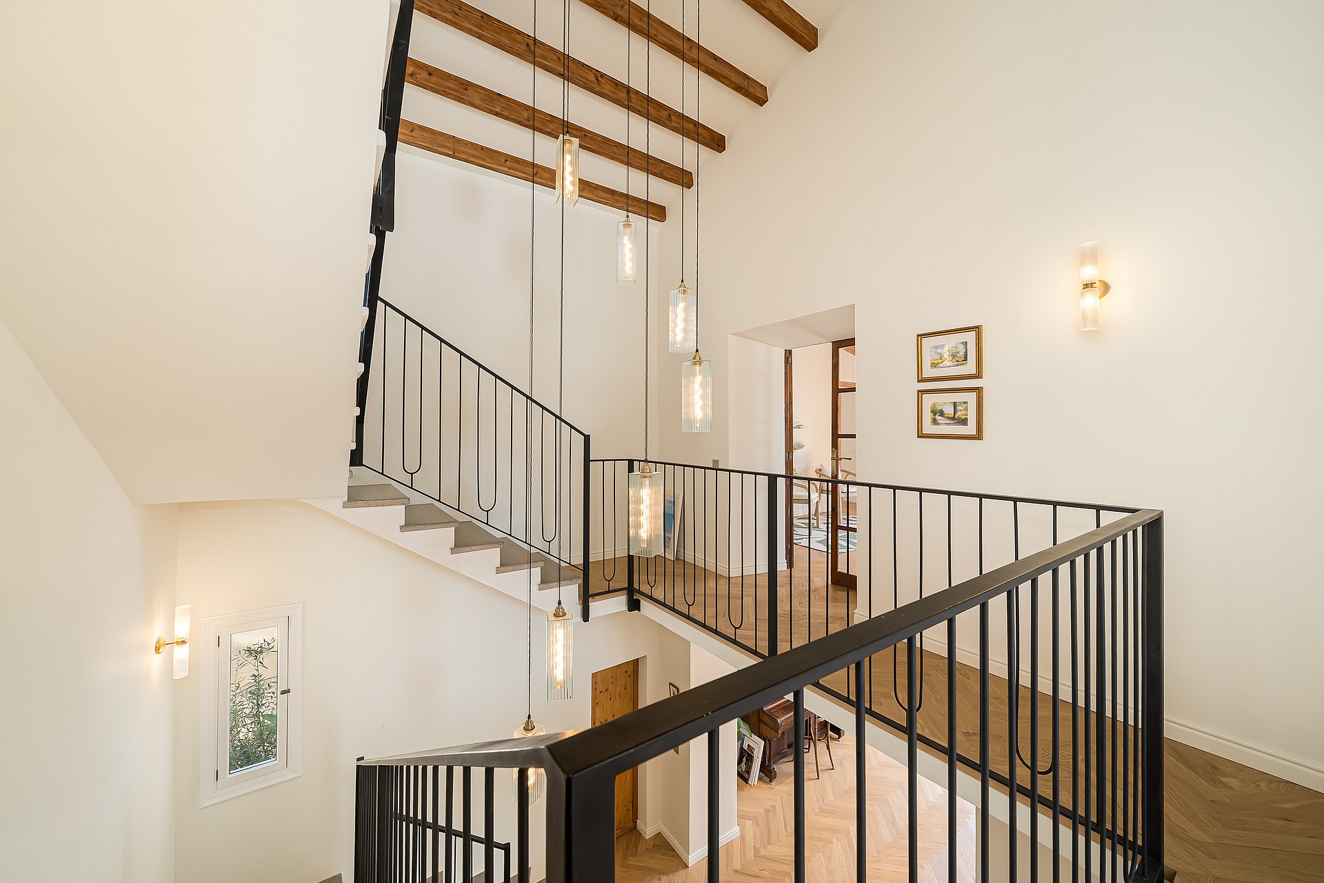 Modern staircase featuring black iron railings, exposed wooden beams, and a cluster of cylindrical glass pendant lights.