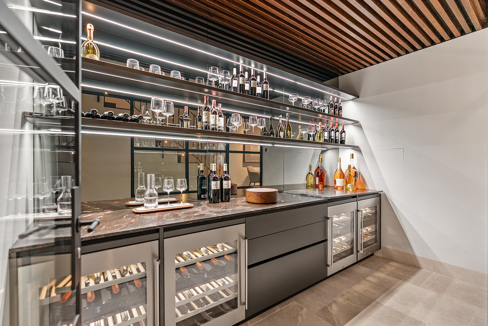 Dark-toned wine cellar with backlit glass shelves, integrated temperature-controlled fridges, and a marble workspace.