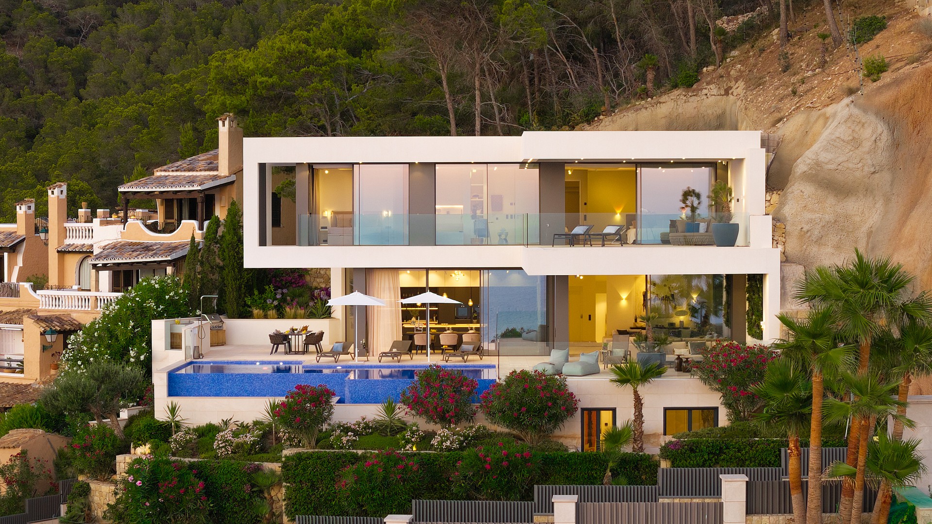 Contemporary white villa built into a hillside, featuring large glass windows, a blue swimming pool, and terraced gardens.