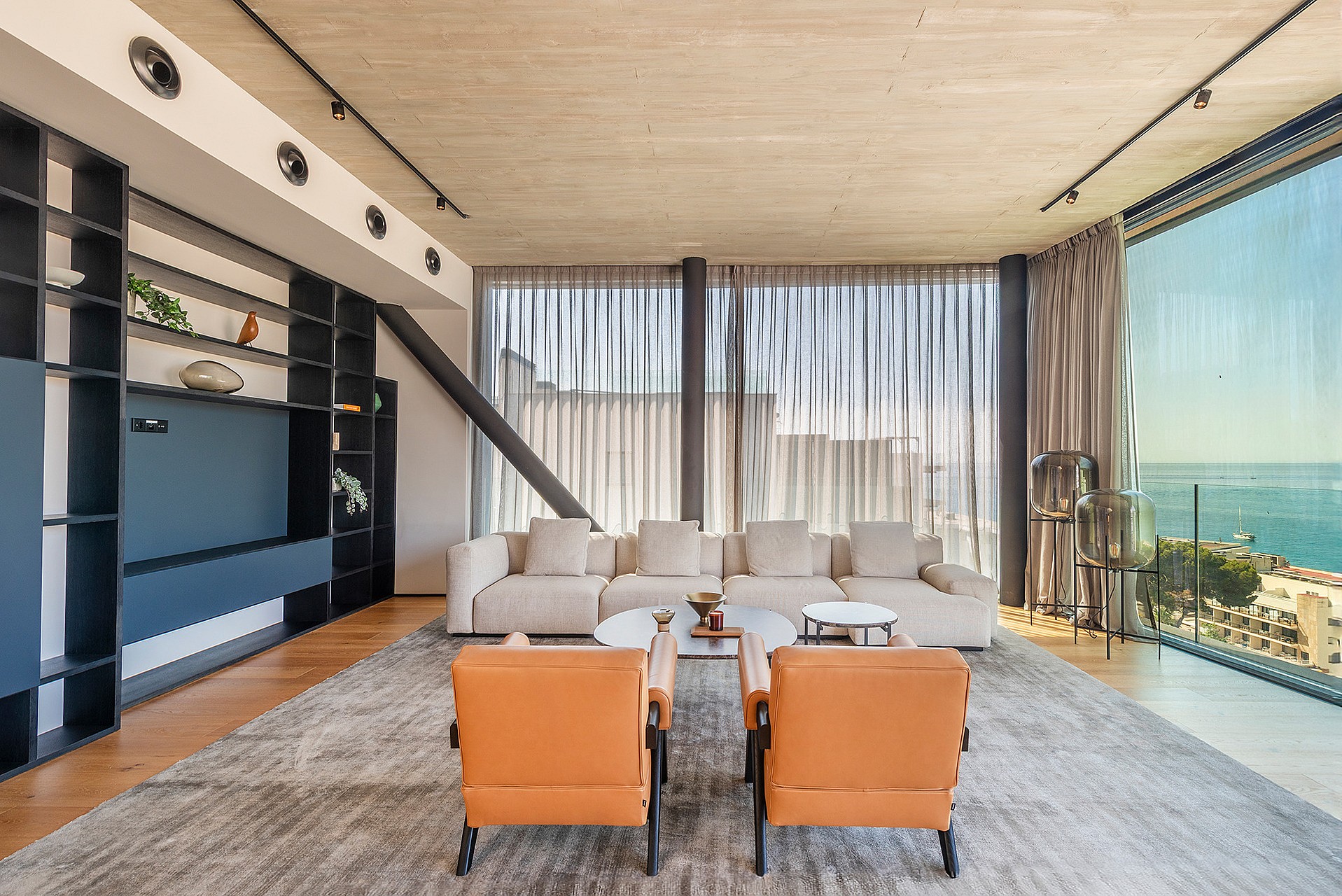 Contemporary living room with a large cream sofa, two orange leather armchairs, and floor-to-ceiling glass overlooking the sea.