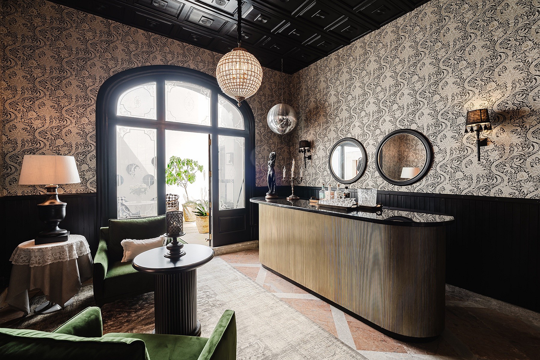 A formal bar area featuring patterned wallpaper, black wainscoting, a gold-toned bar counter, and velvet armchairs near a large arched window.
