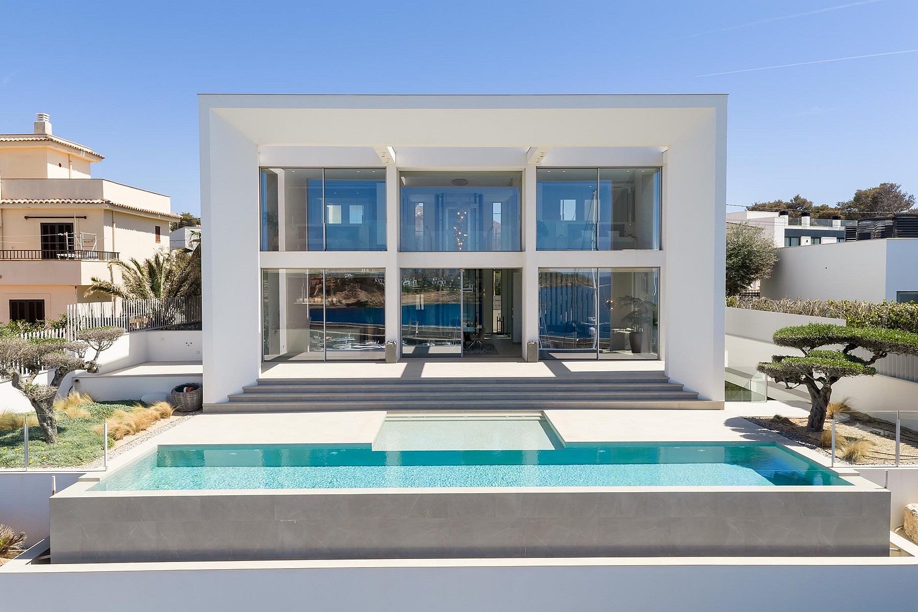 Two-story white modern villa with floor-to-ceiling glass walls, a rectangular swimming pool, and stone-paved terrace.
