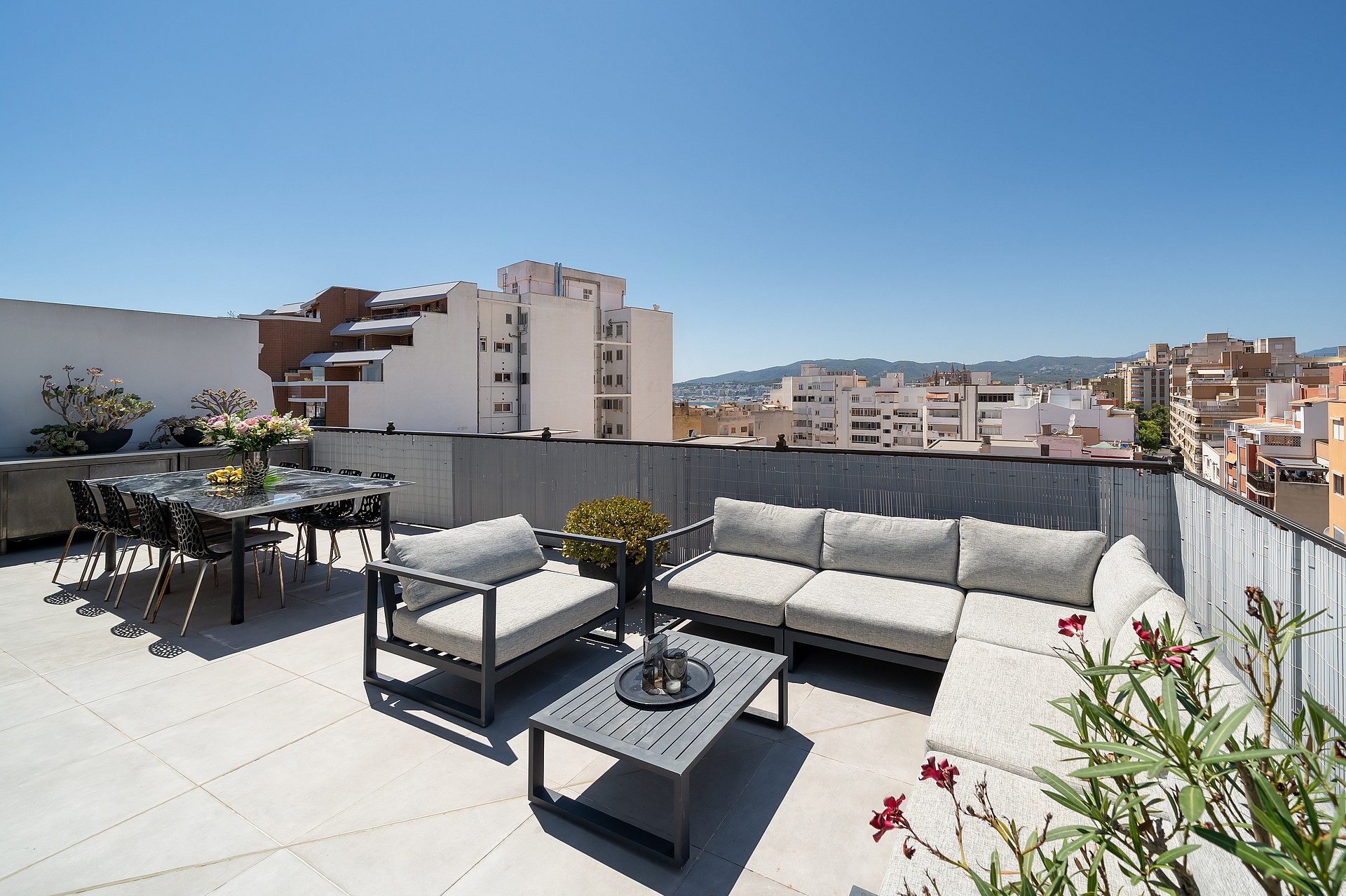 A spacious rooftop terrace featuring grey modular outdoor furniture, a glass dining table, and wide views of the city skyline.