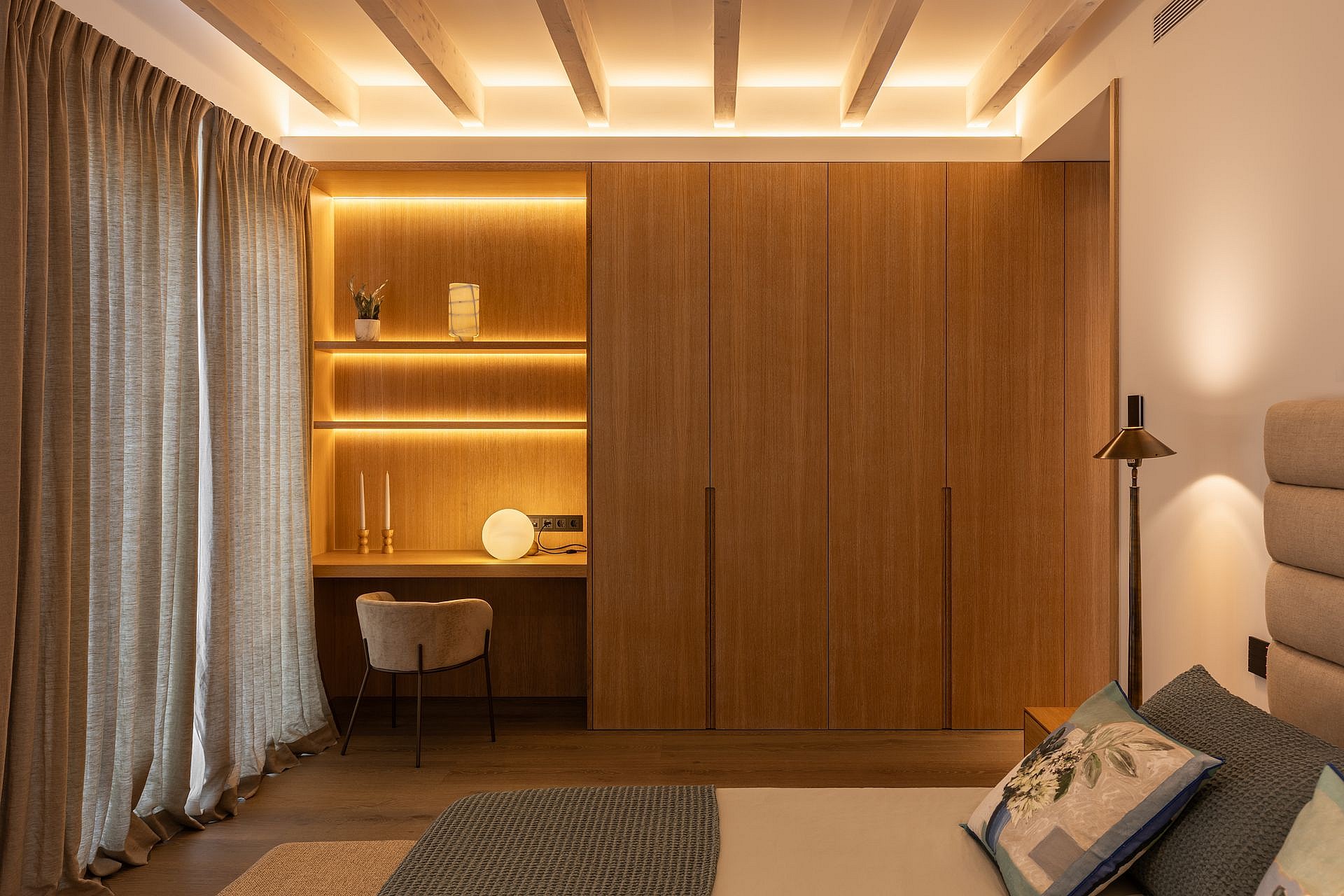 Bedroom featuring custom floor-to-ceiling wooden wardrobe, built-in shelving desk area, and neutral fabric curtains.