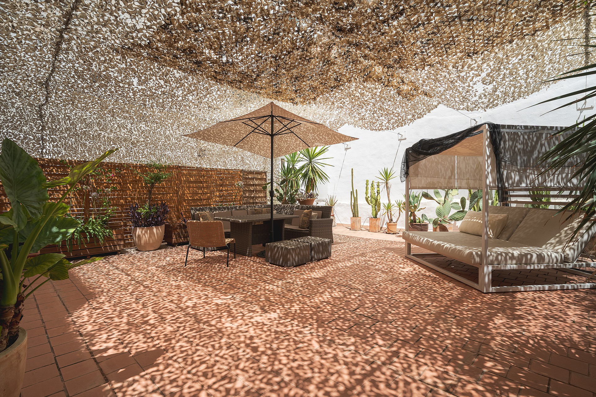 Outdoor terrace with camouflage netting shade, dining table, and a daybed on a terracotta tiled floor.