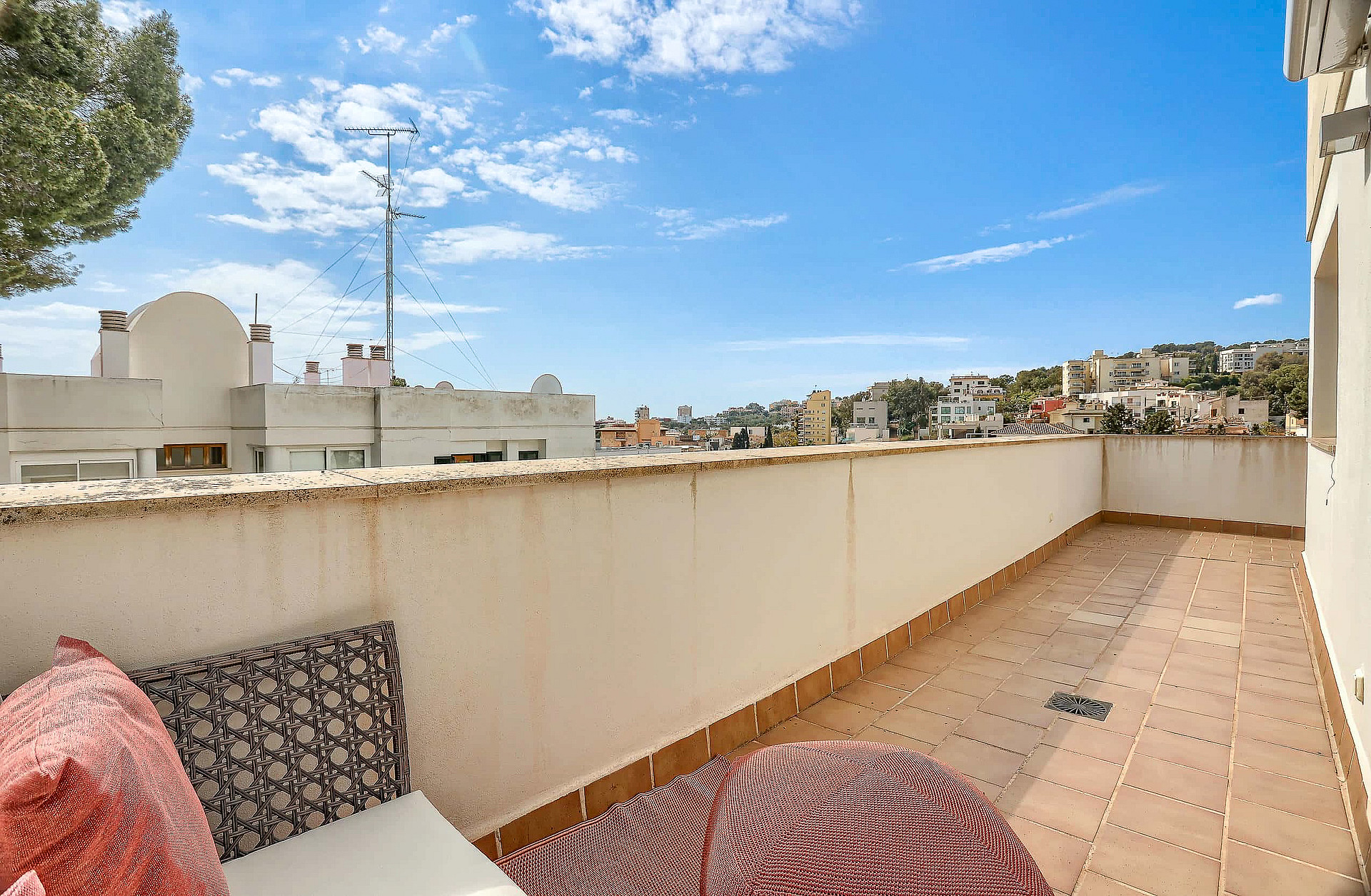 A private outdoor terrace with tiled flooring, a low perimeter wall, and patterned cushions under a bright blue sky.