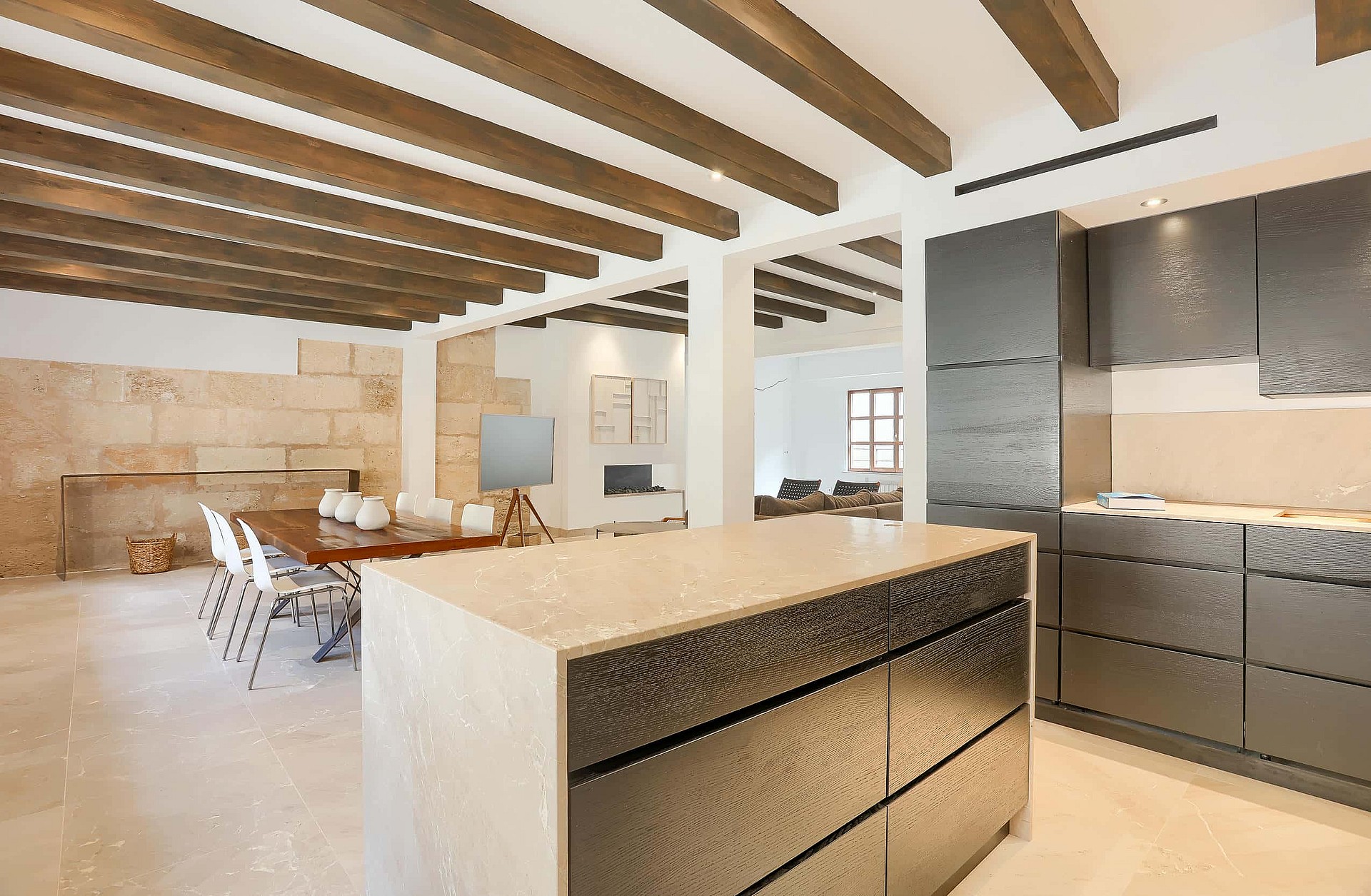 A modern kitchen with a dark central island and a dining table in an open-plan room featuring exposed ceiling beams and limestone walls.