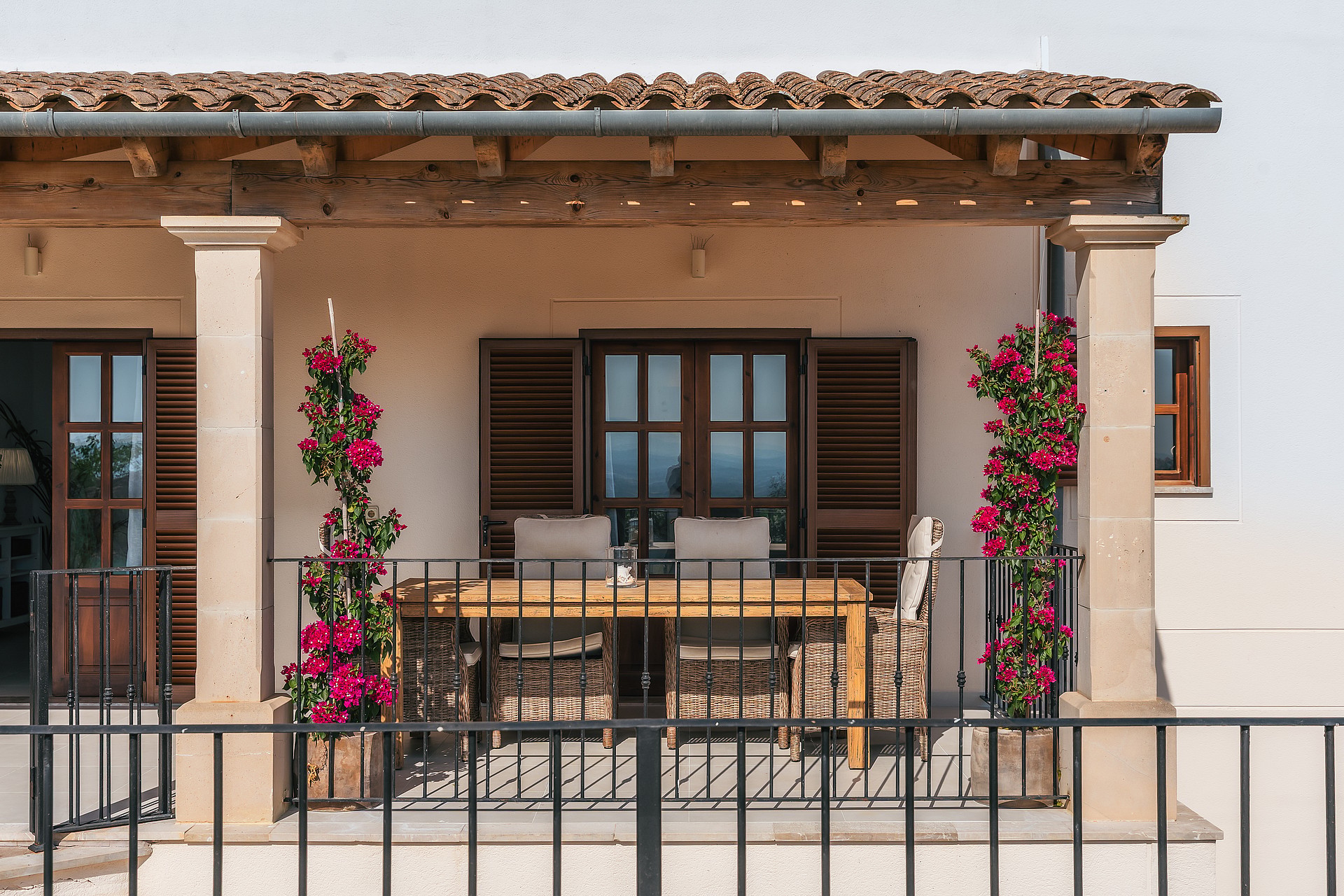 Porche con columnas de piedra, mesa de comedor de madera, contraventanas oscuras y buganvillas en flor.