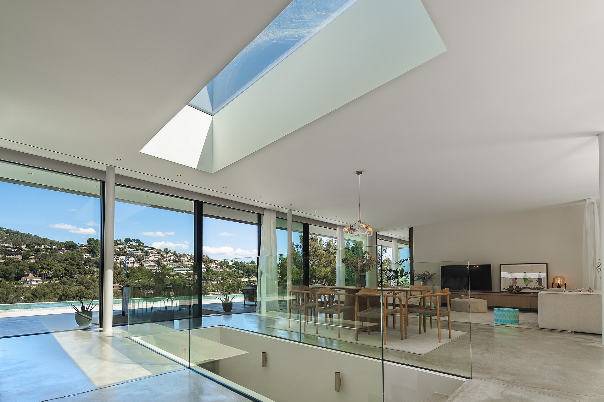 Modern living and dining room featuring floor-to-ceiling glass walls, a large skylight, and views of Son Vida hills.