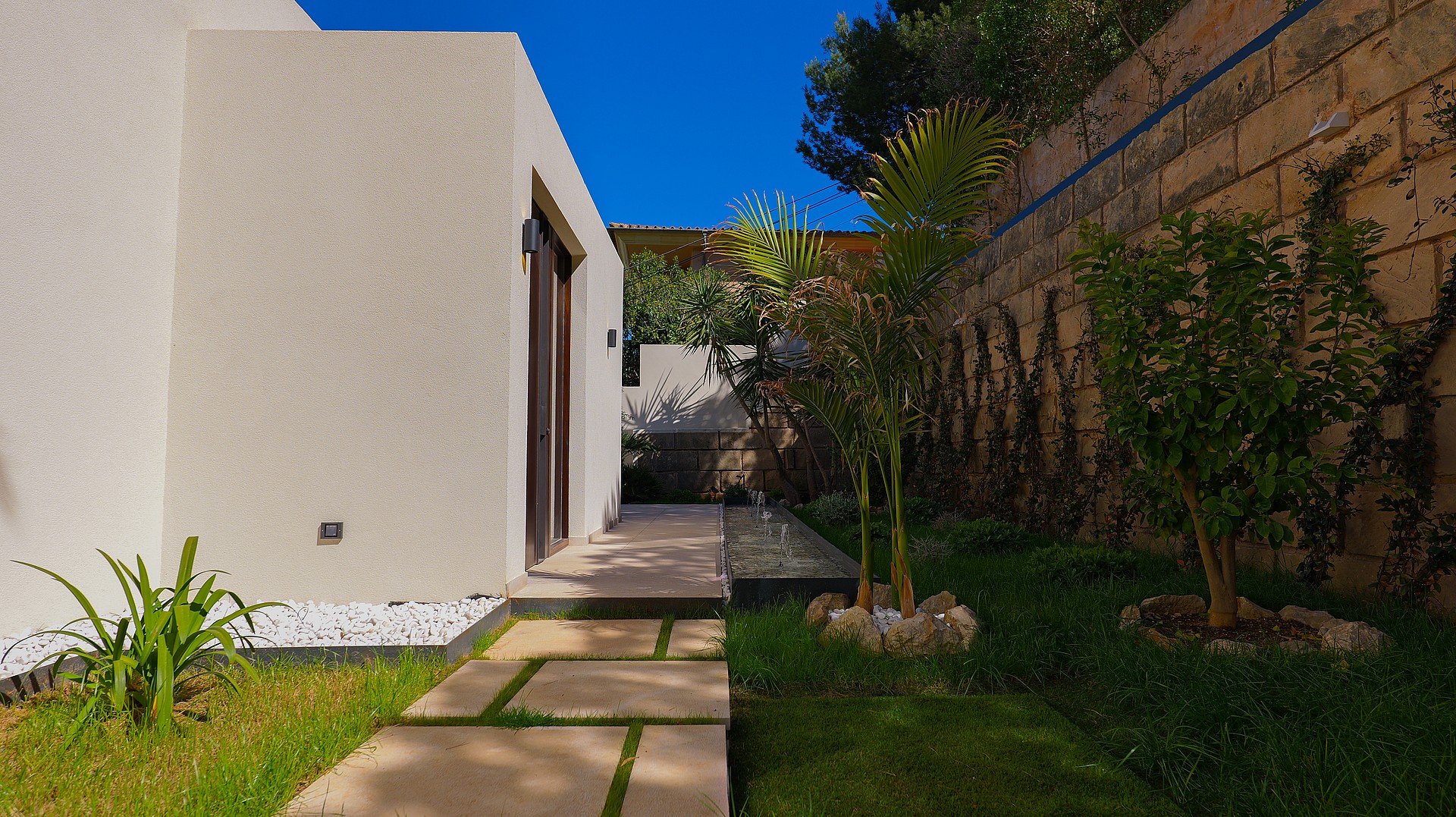 A minimalist white exterior wall with a dark framed doorway and a stone slab pathway leading through a green garden.