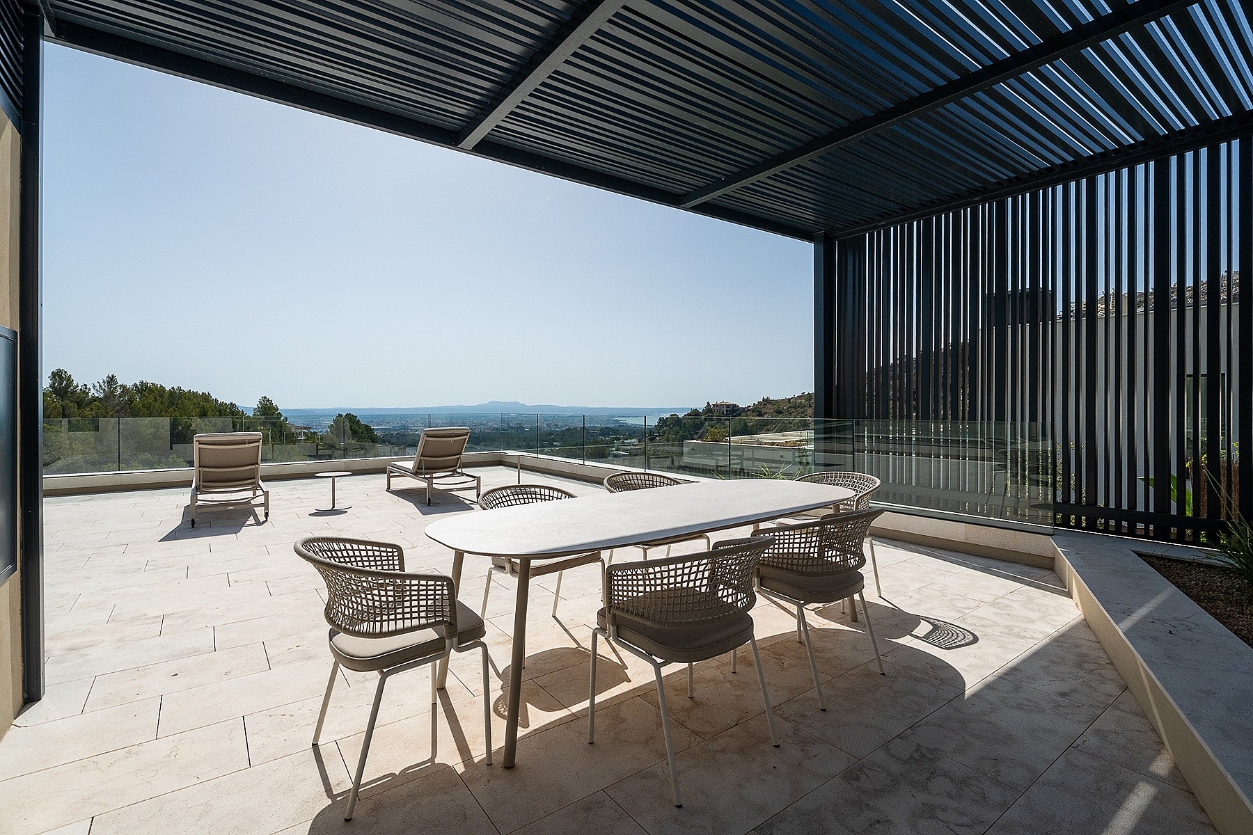 A wide outdoor terrace featuring a white stone floor, a dining table with woven chairs, and a black slatted pergola roof.