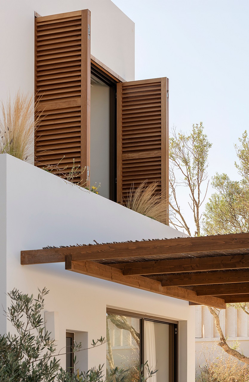 Exterior detail of a white building featuring dark wood shutters and a rustic timber pergola against a clear sky.