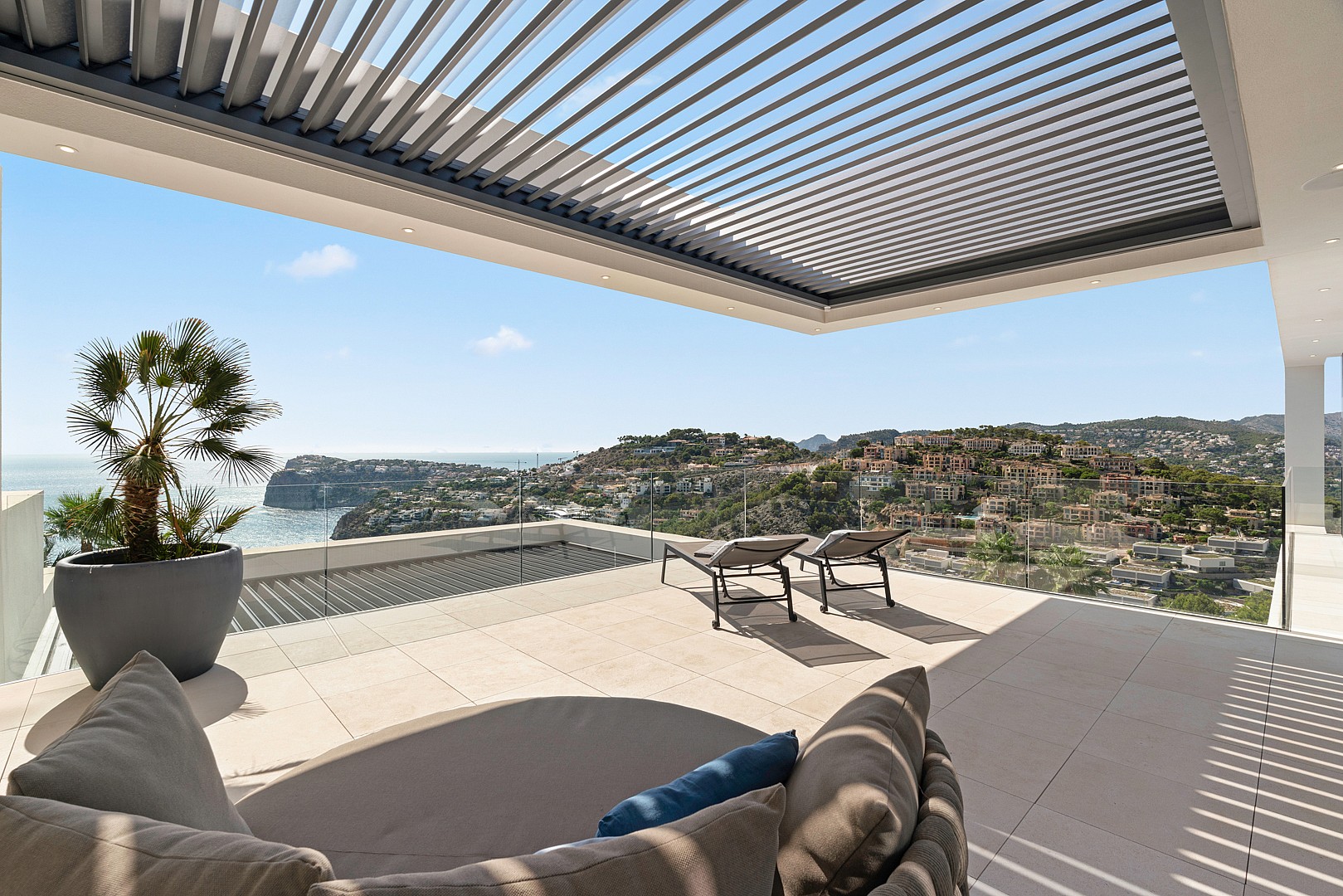 A covered terrace with a louvered pergola and lounge seating overlooking the sea and hillside villas in Port d'Andratx.