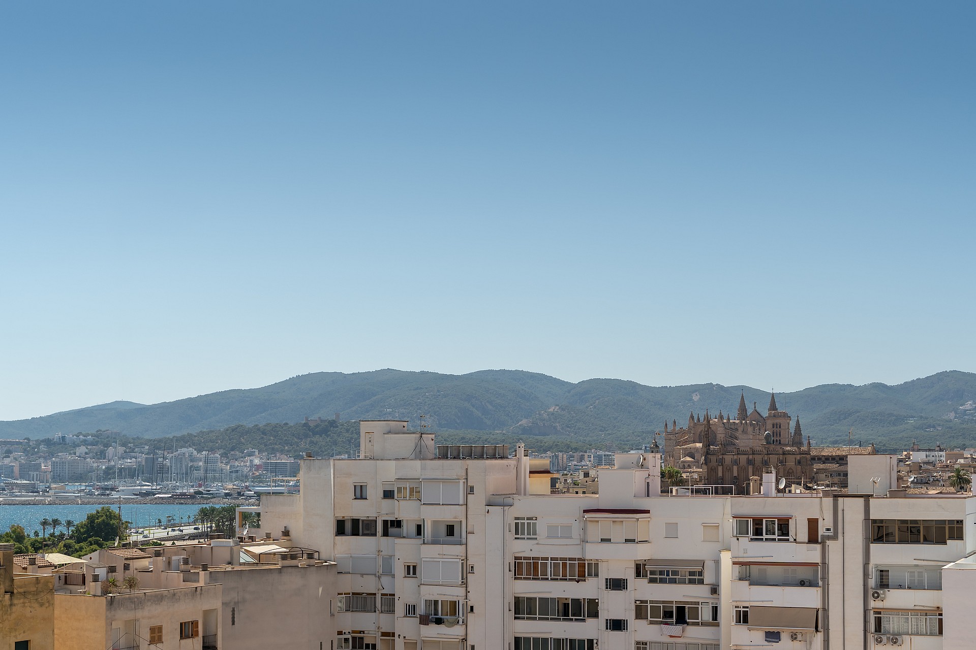 Panoramic view of a coastal city with white buildings and distant mountains under a clear blue sky.