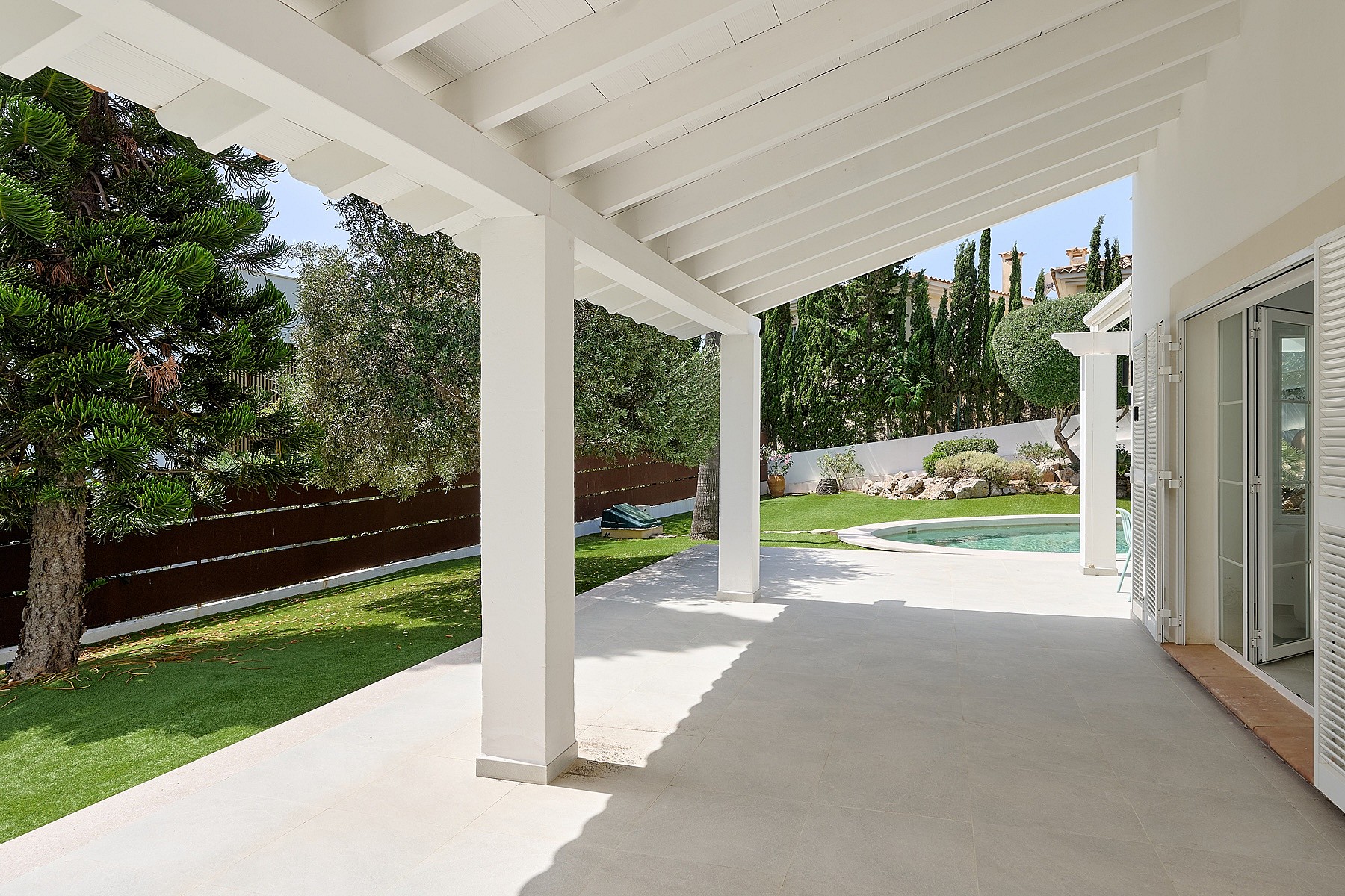 Terraza cubierta blanca con vigas de madera frente a césped artificial, piscina circular, pinos y cipreses