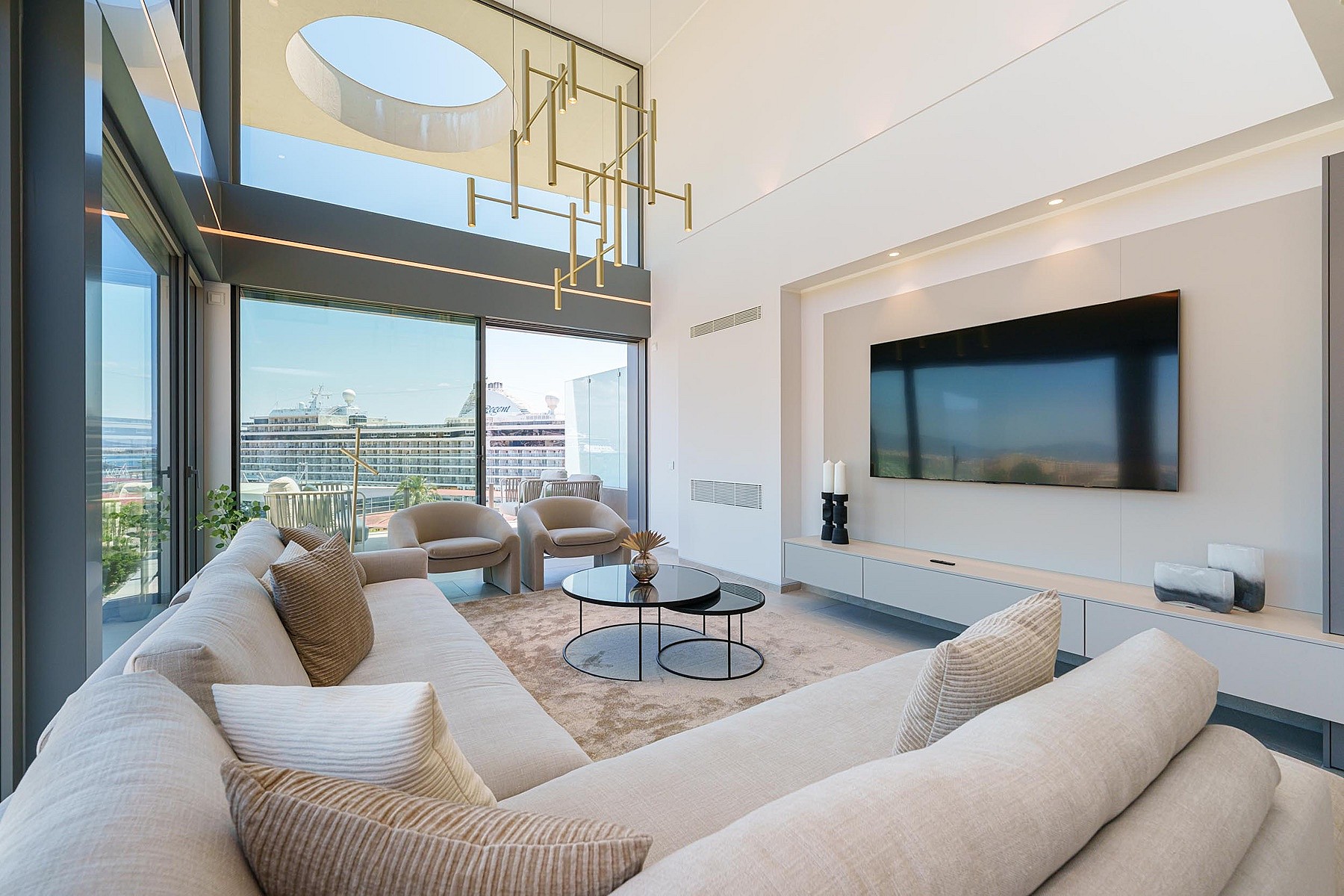 Salón moderno de doble altura con sofá, mesas auxiliares circulares y ventanales de cristal orientados al mar.